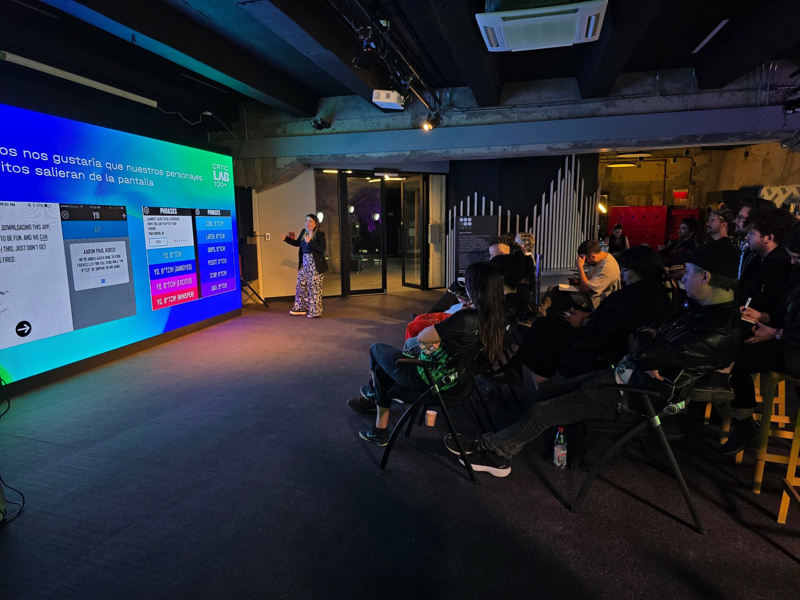 Presenter standing in front of a large screen displaying colorful slides, audience seated in a dark room, some taking notes.