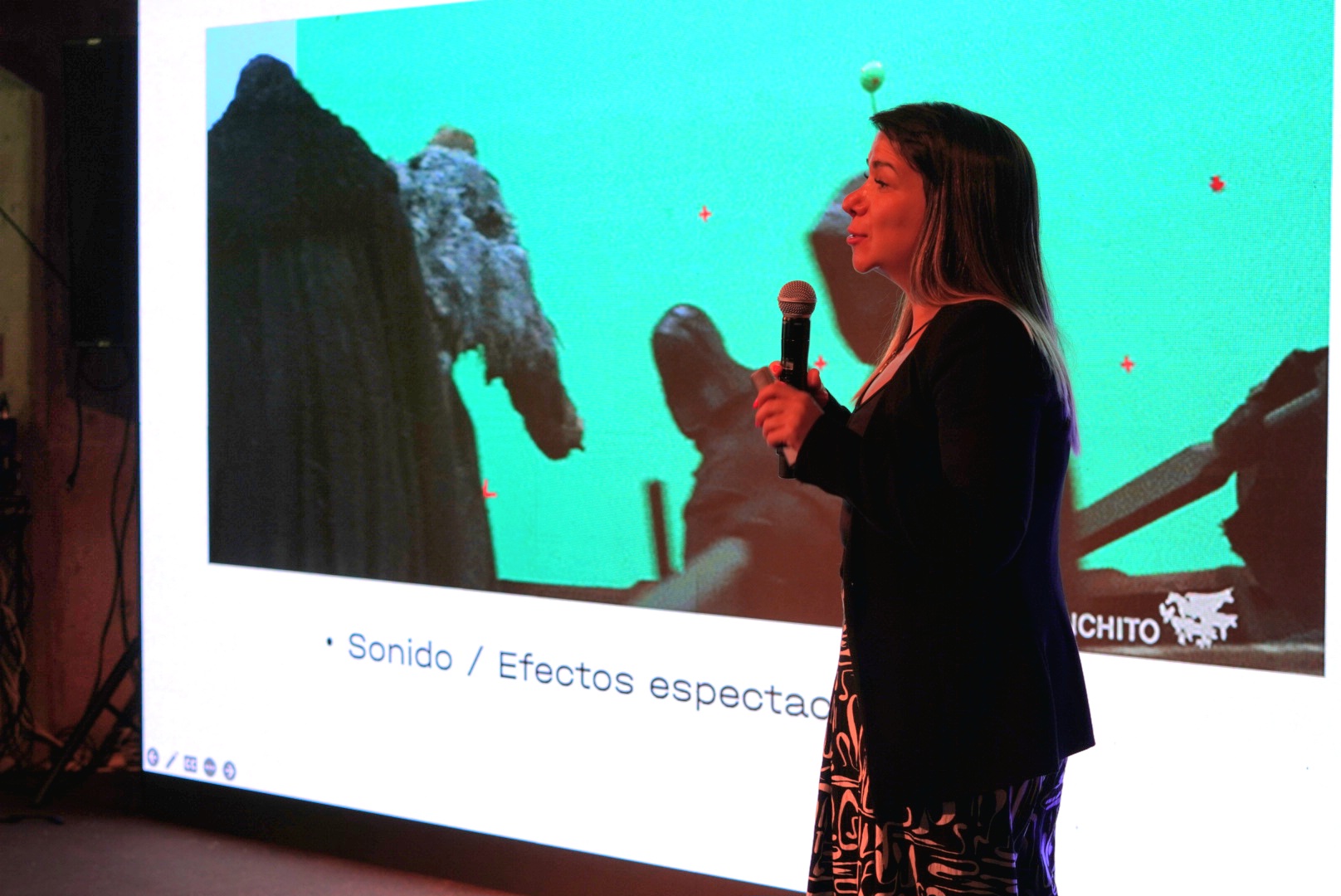 Woman holding a microphone presents in front of a large screen with a blue background and mountain images.