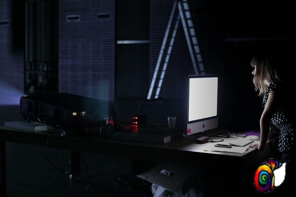 Person working at a desk with a computer monitor in a dark room, illuminated by the screen, with a projector nearby.