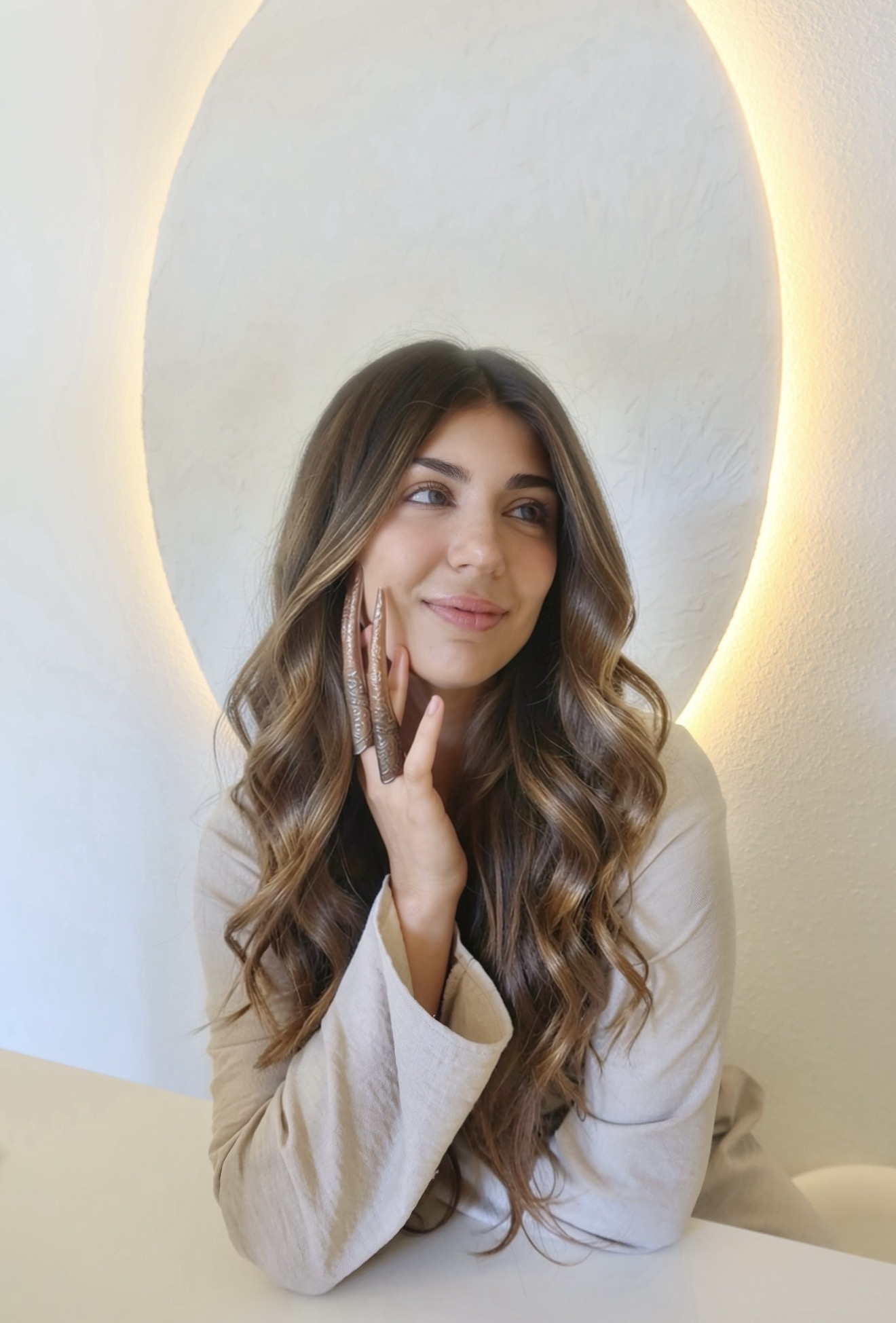 Young woman with long wavy hair smiling, resting her face on her hand, in front of a backlit oval mirror.