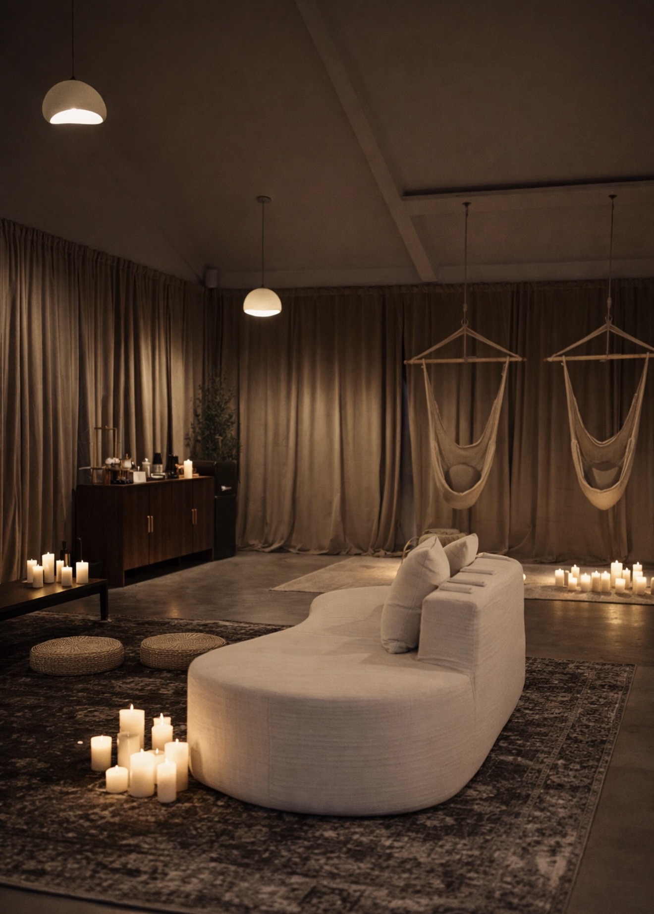 Living room with hanging chairs, candles, and a curved sofa, dimly lit with warm lighting and curtains in the background.
