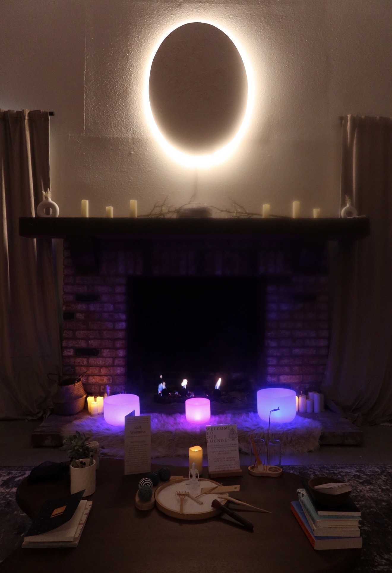 Decorative fireplace with candles and glowing objects, illuminated by a bright oval light on the wall above, in a cozy room.