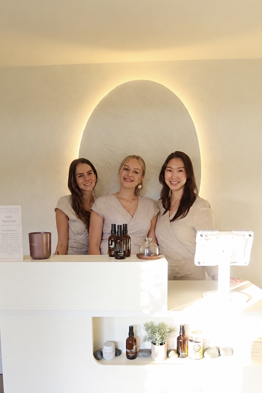 Three women standing behind a white counter with bottles and plants, smiling, in a well-lit room with a circular wall feature.