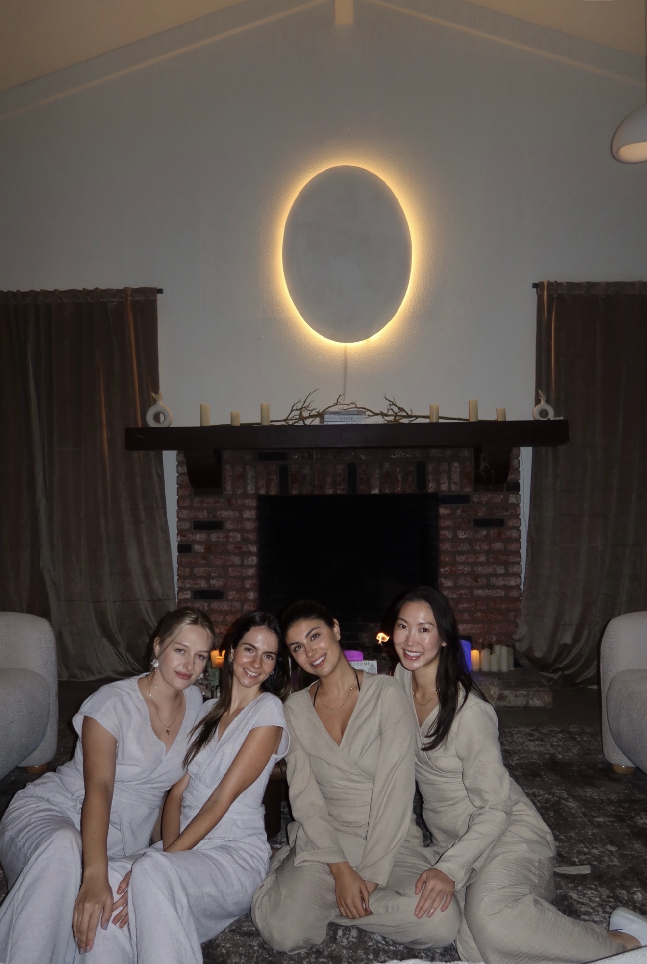 Four women sit on the floor in front of a brick fireplace with a glowing oval light above. Two curtains frame the fireplace.