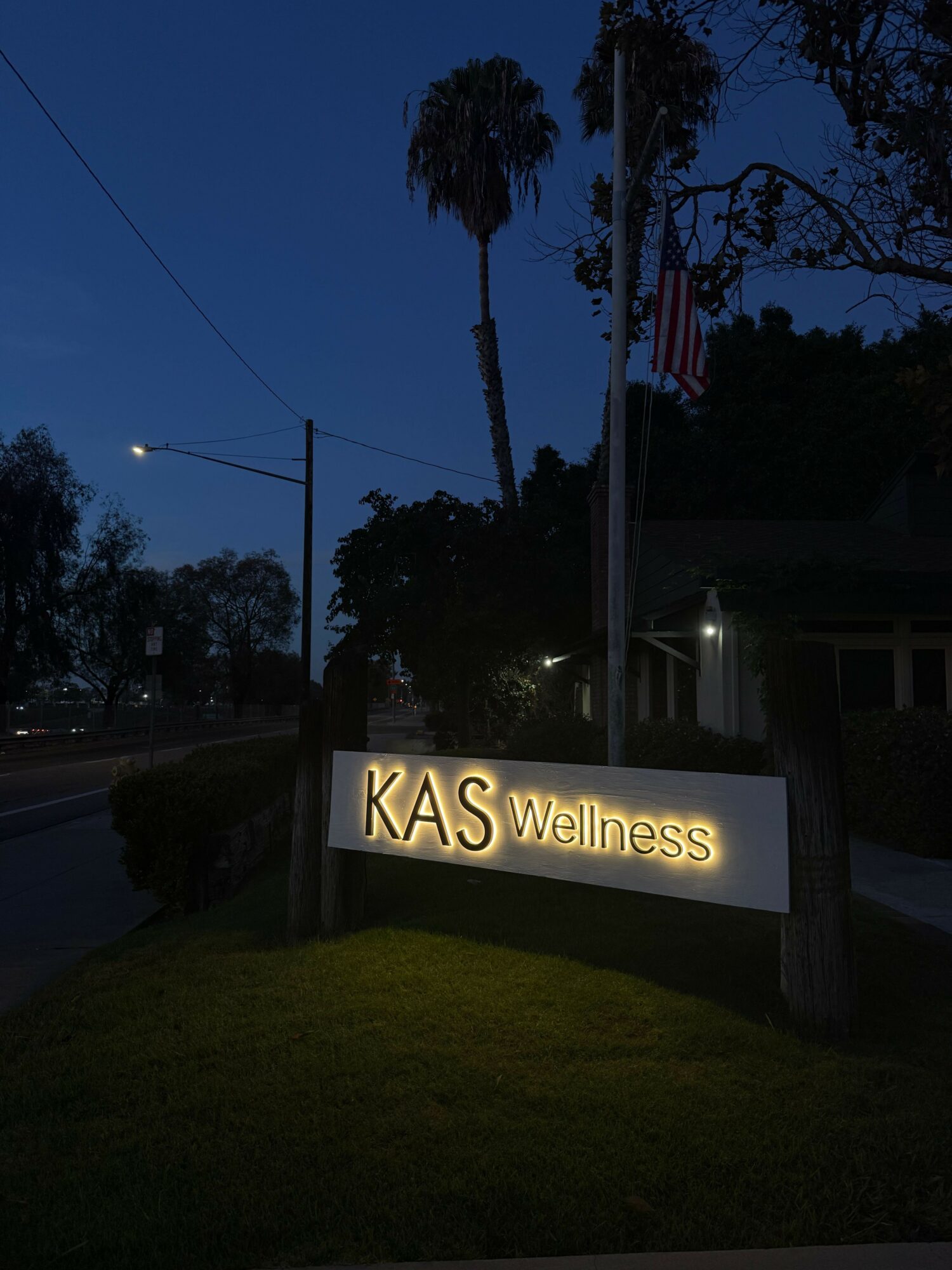 Illuminated sign reads 'KAS Wellness' on grass, with trees and a flagpole in the background during dusk.