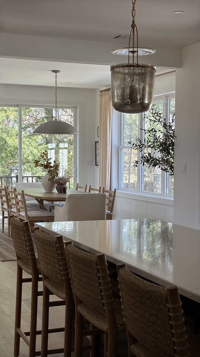 Dining area with a large table, chairs, and a hanging light fixture, with windows showing trees outside.