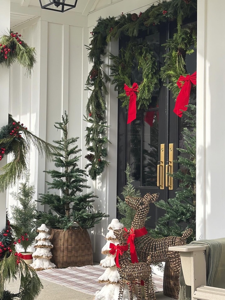 Decorated front door with Christmas wreath, red bows, small Christmas trees, and festive decorations outside a white house.