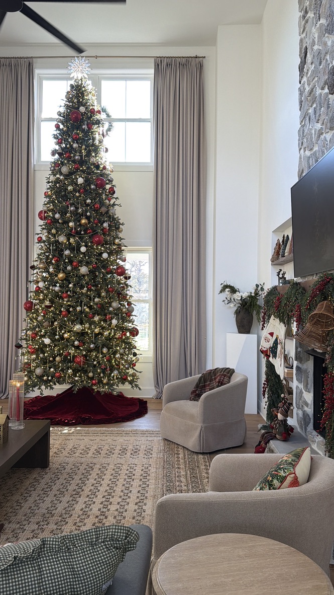 Tall decorated Christmas tree near window in a living room with armchairs and fireplace.