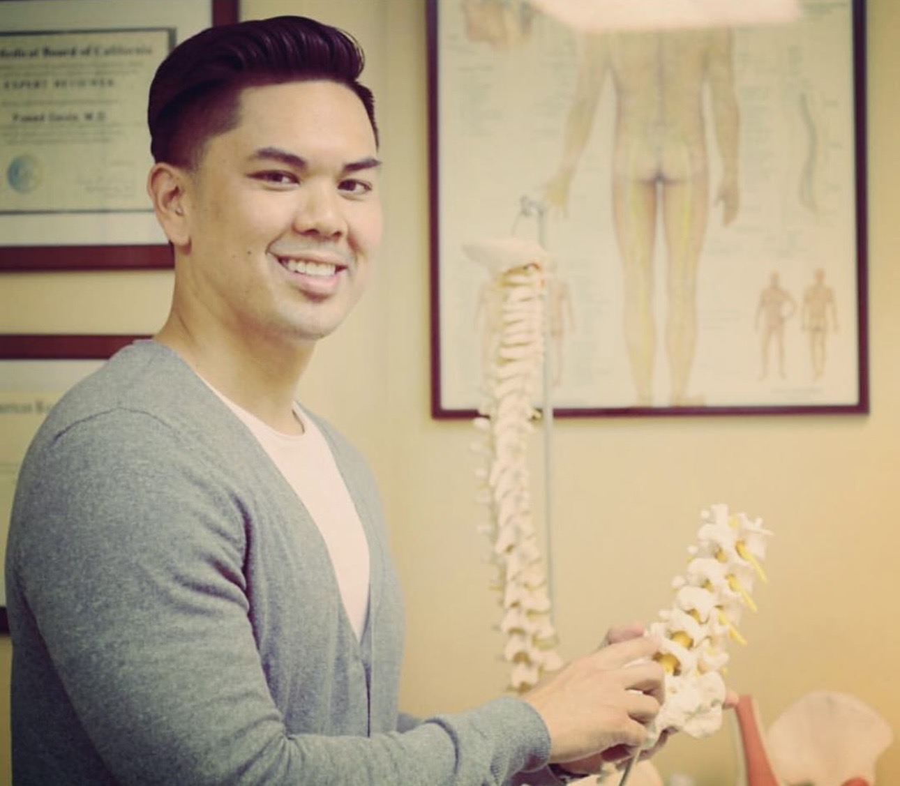 Young man smiling in medical office holding a model of a human spine, with anatomical posters on the wall.