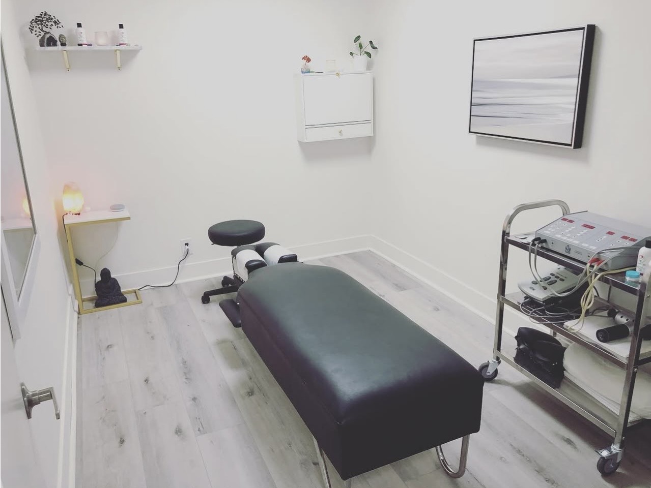 Medical examination room with a black examination table, rolling cart, wall-mounted monitor, and shelves, all in a white room.