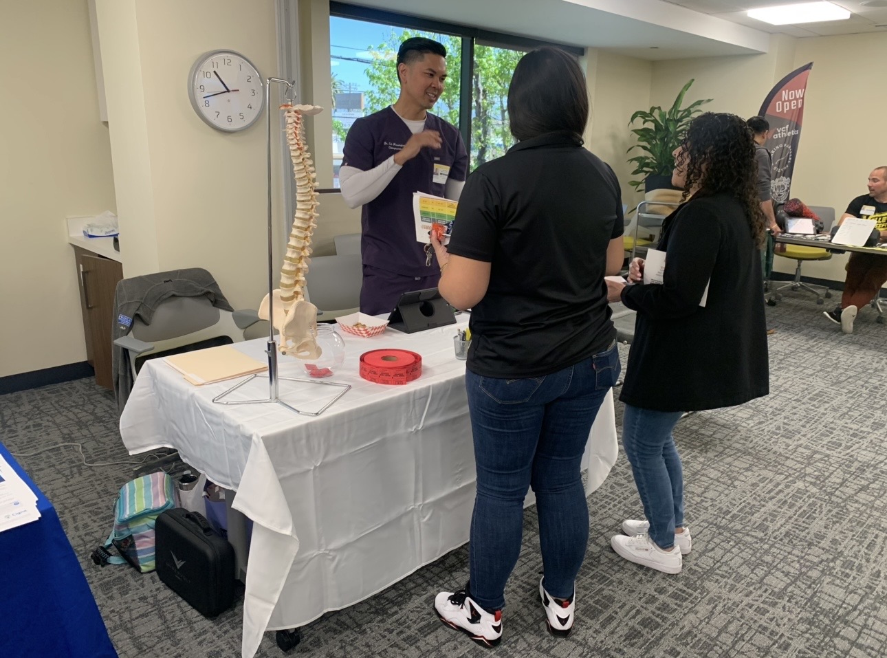 Person explaining a model of the human spine to three people at an indoor event, with a table and clock in background.