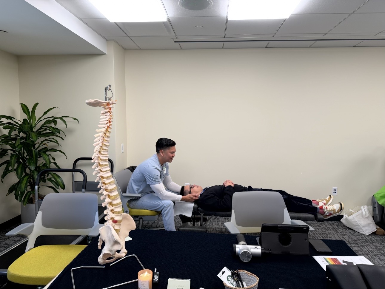 Person lying on a table with a healthcare professional holding their hand in a room with chairs, a plant, and a model of a human spine.