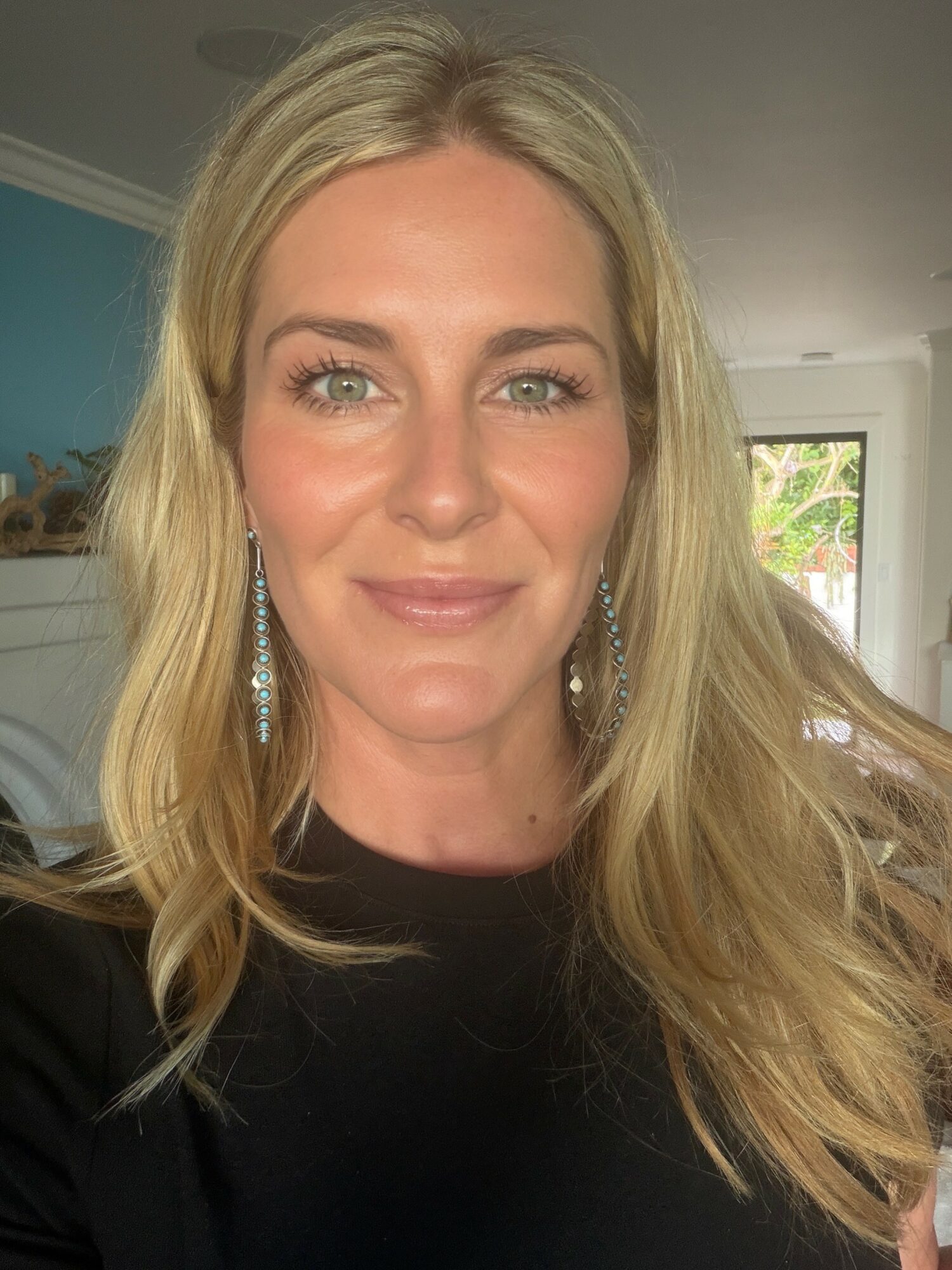 Portrait of a woman with long blonde hair, smiling, wearing earrings, indoors with a window in the background.