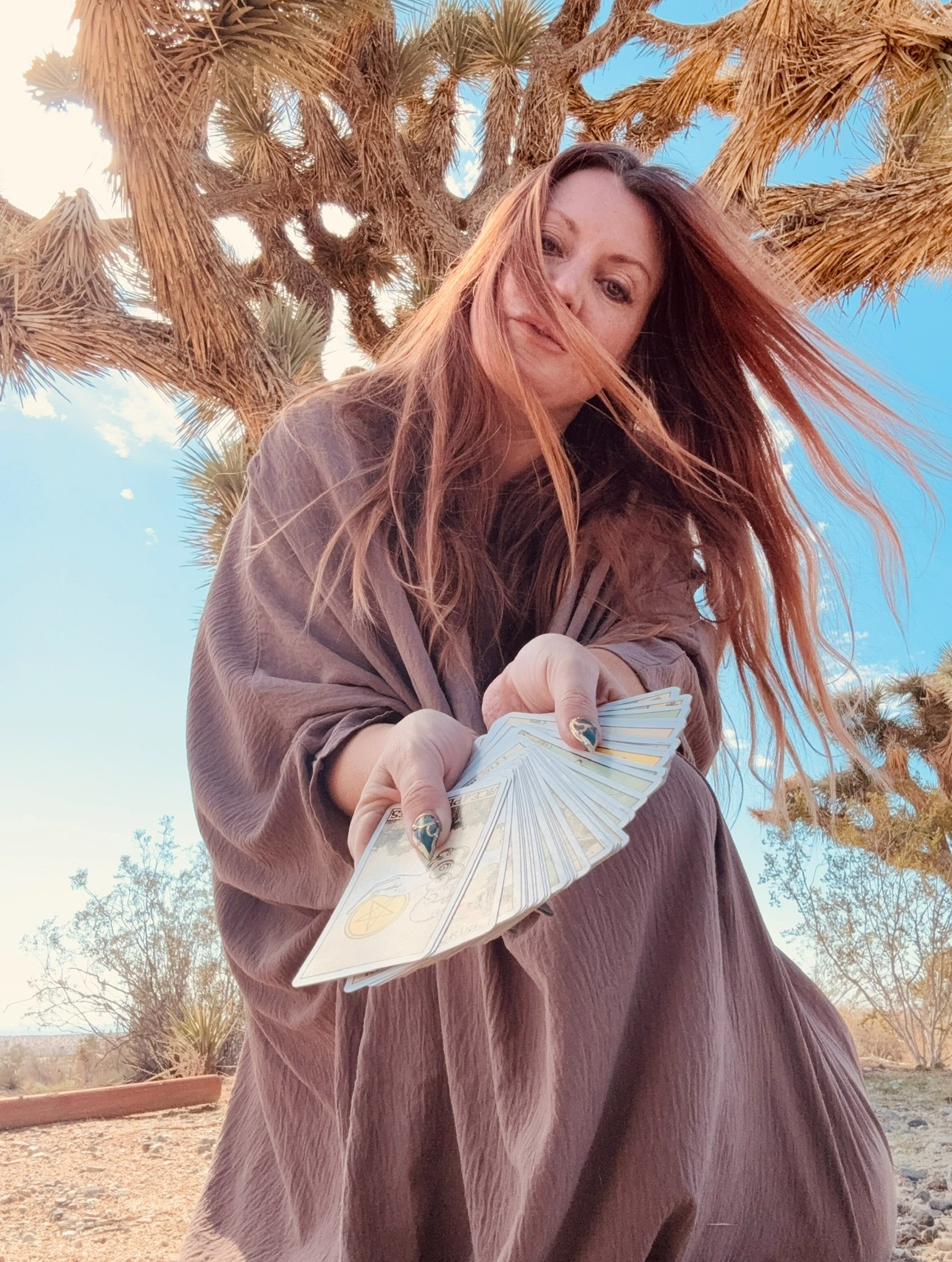 Woman with long hair holding a fan outdoors, desert landscape with a large tree and blue sky behind her.