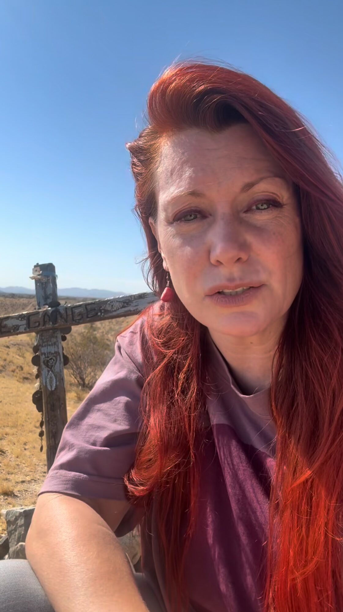 Woman with red hair outdoors, blue sky background, leaning on a wooden fence, looking at the camera.