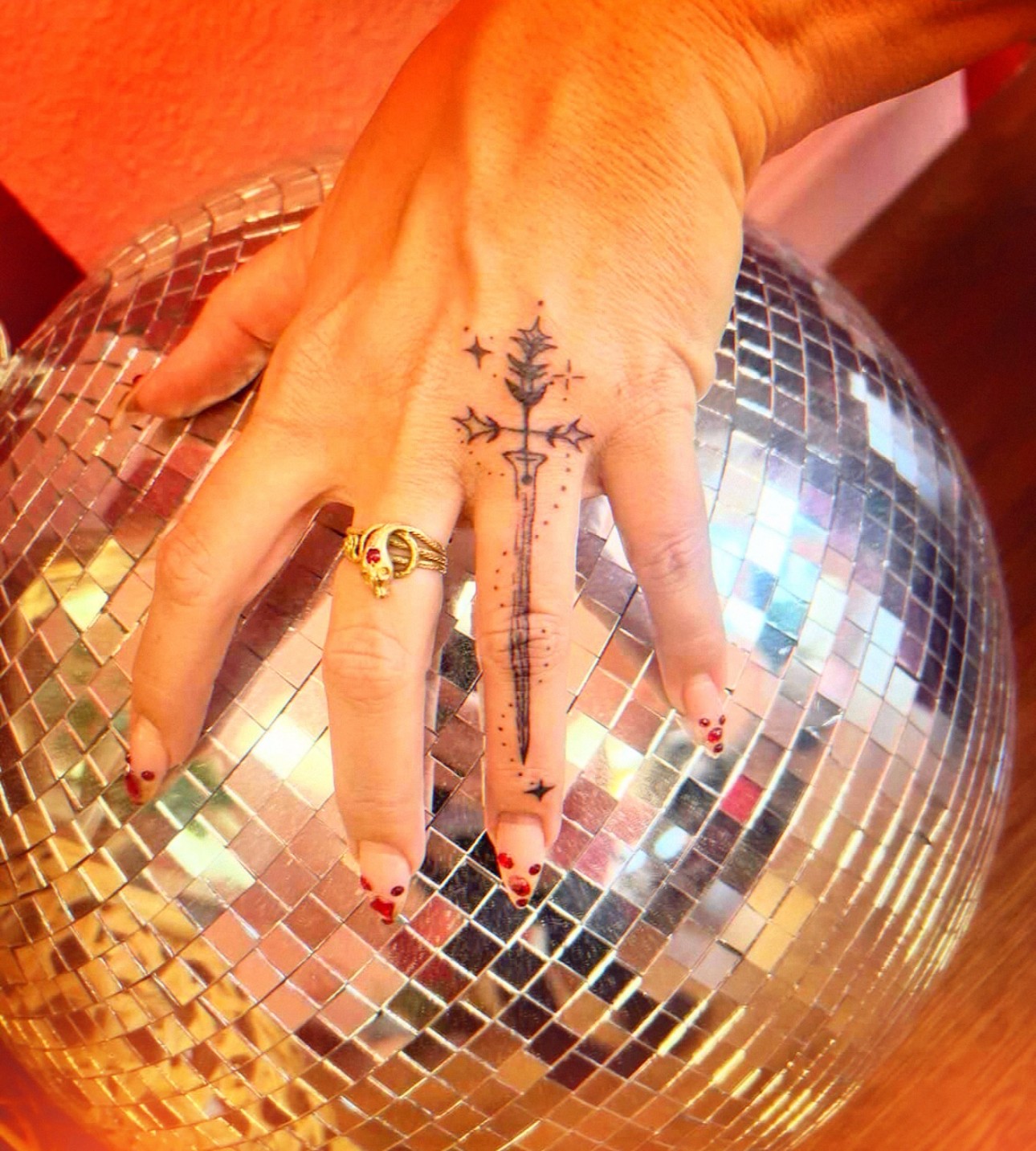 Hand with a tattoo resting on a disco ball, wearing a ring, with red nail polish, against an orange background.