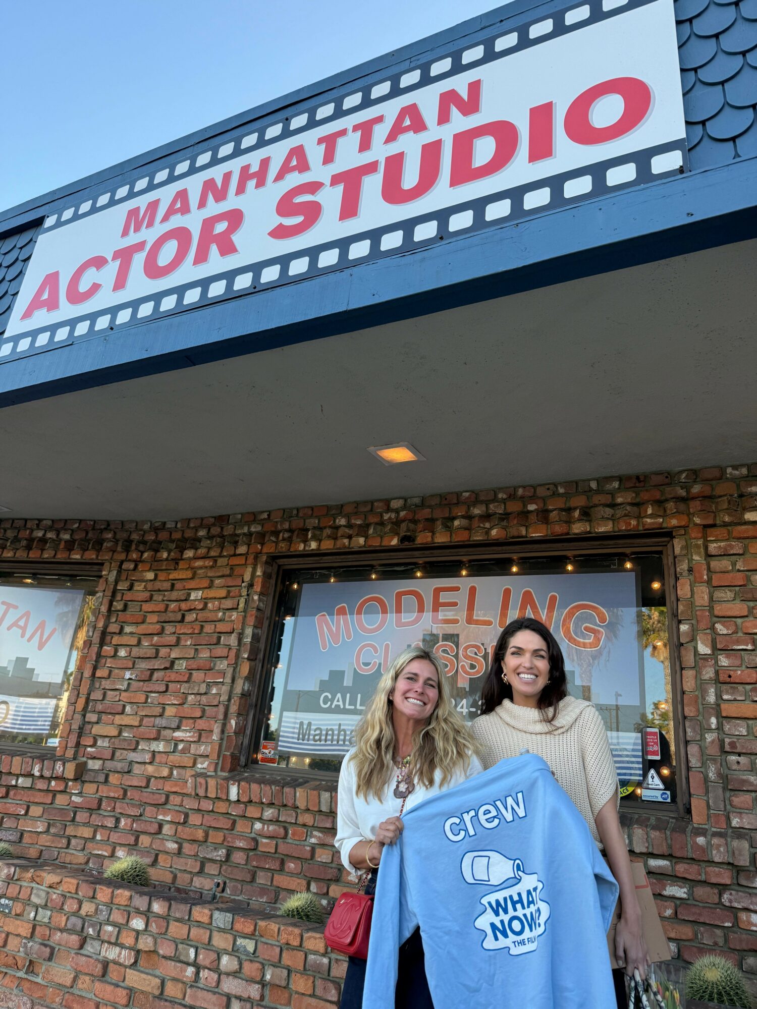Two women stand outside a building with a brick wall and a sign for Manhattan Acting Studio, smiling at the camera.
