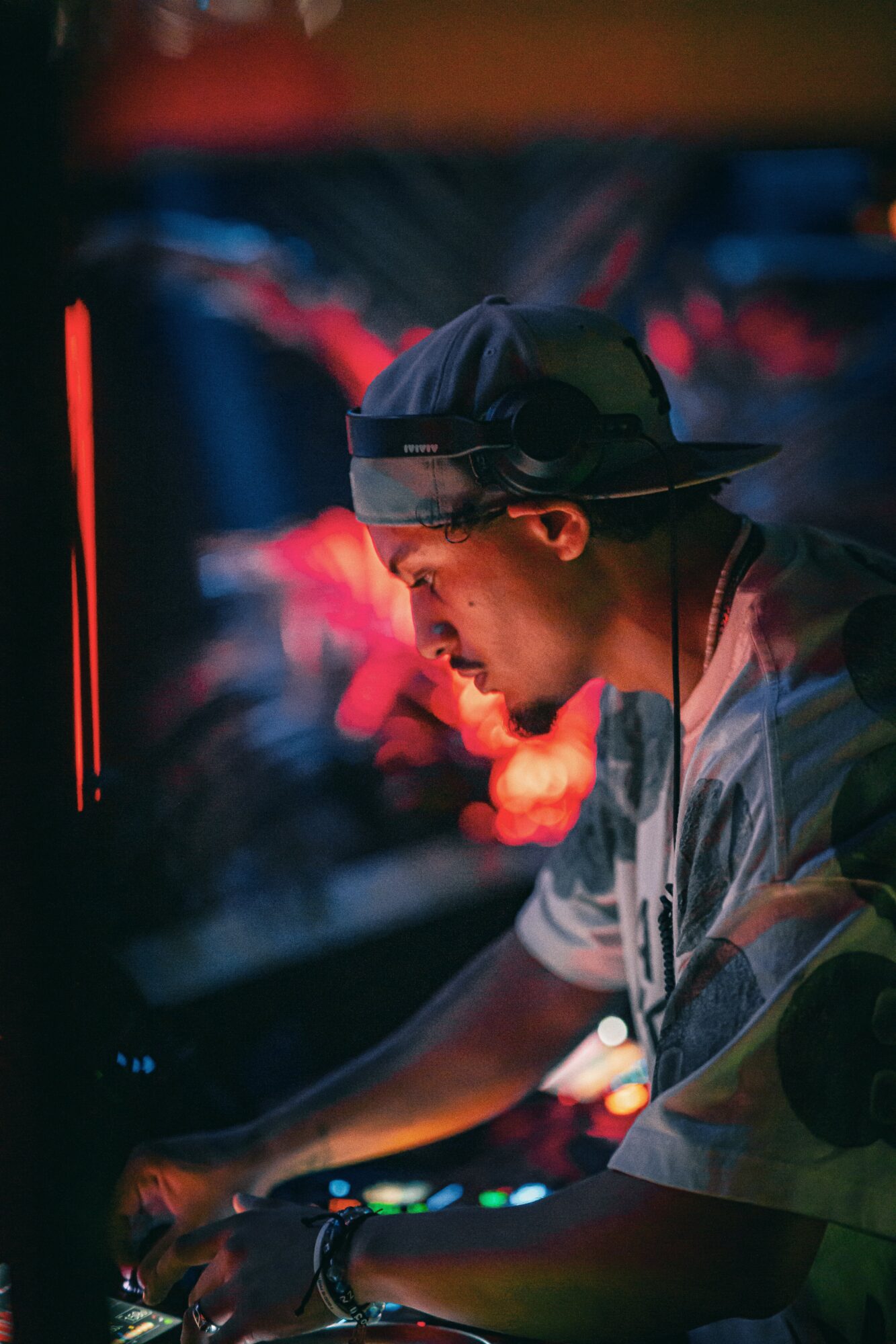 DJ wearing a cap and glasses, focused on mixing music in a dimly lit environment with colorful lights.