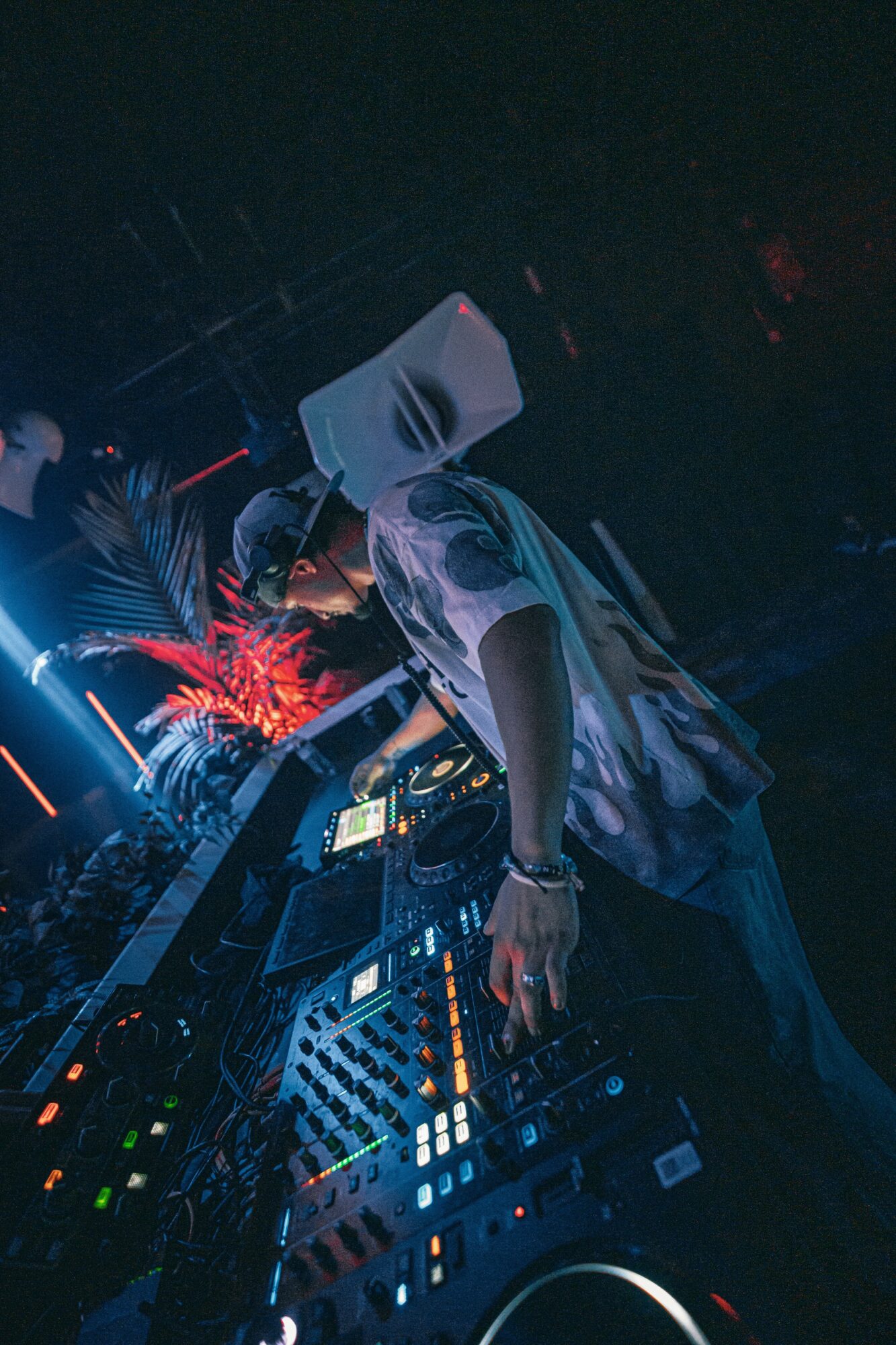 DJ wearing a patterned shirt operates equipment in a dark club with colorful lights and a plant in the background.
