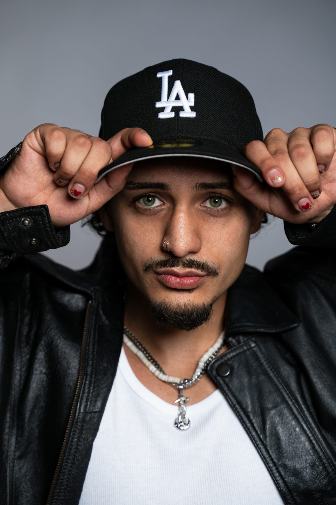 Young man with a mustache and goatee wearing a black LA cap, black leather jacket, and white shirt, holding the cap's brim.