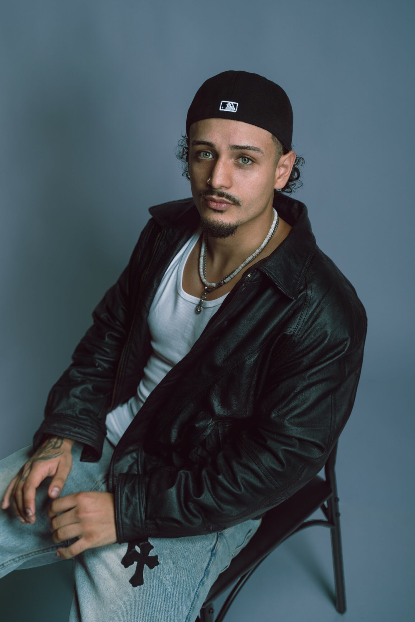 Young man with a black cap, leather jacket, white shirt, and jewelry, sitting on a chair against a blue background.