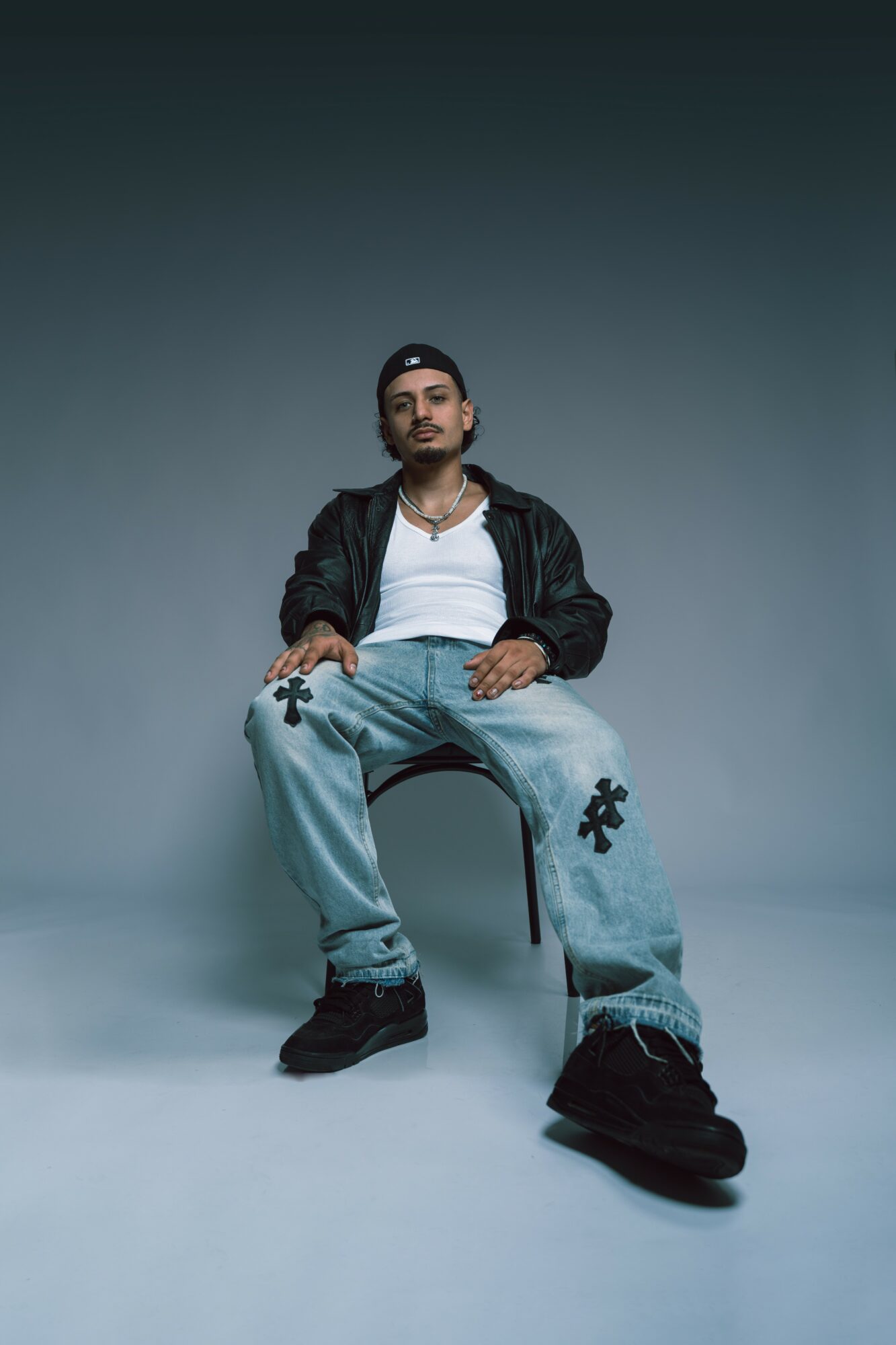 Young man sitting on a chair against a plain background, wearing a black jacket, white shirt, and light blue jeans.