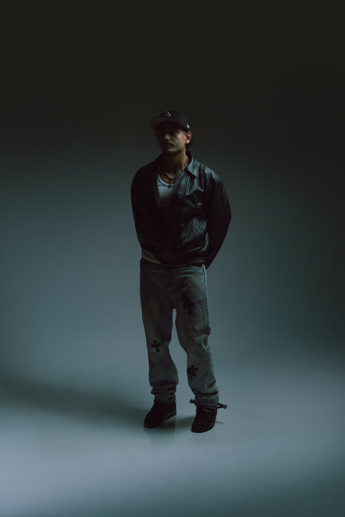 Person standing in a dark, gradient background, wearing a jacket, pants, and a cap, with hands in pockets.