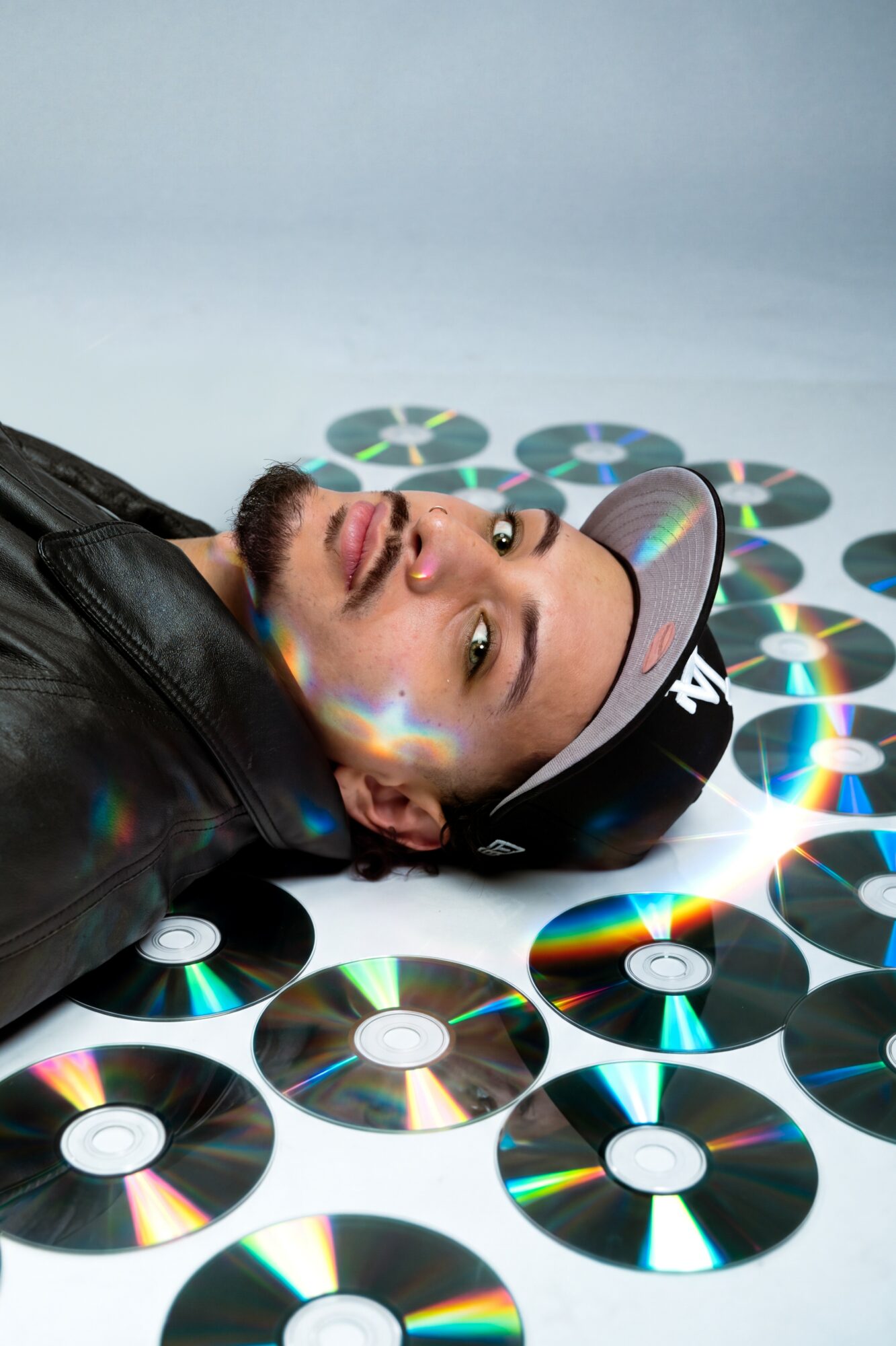 Person lying on the floor surrounded by CDs, wearing a cap and leather jacket, looking up at the camera.