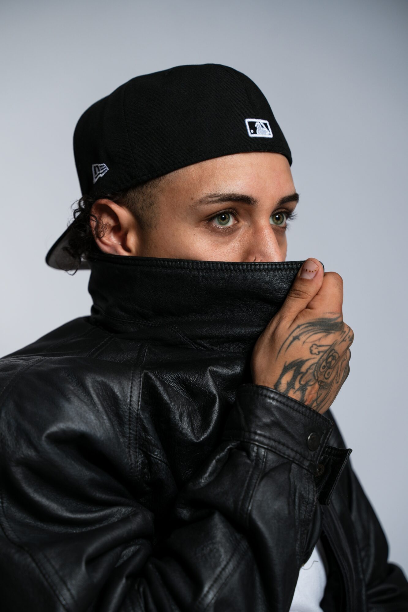 Person wearing a black cap and leather jacket, partially covering their face with the collar, against a plain background.
