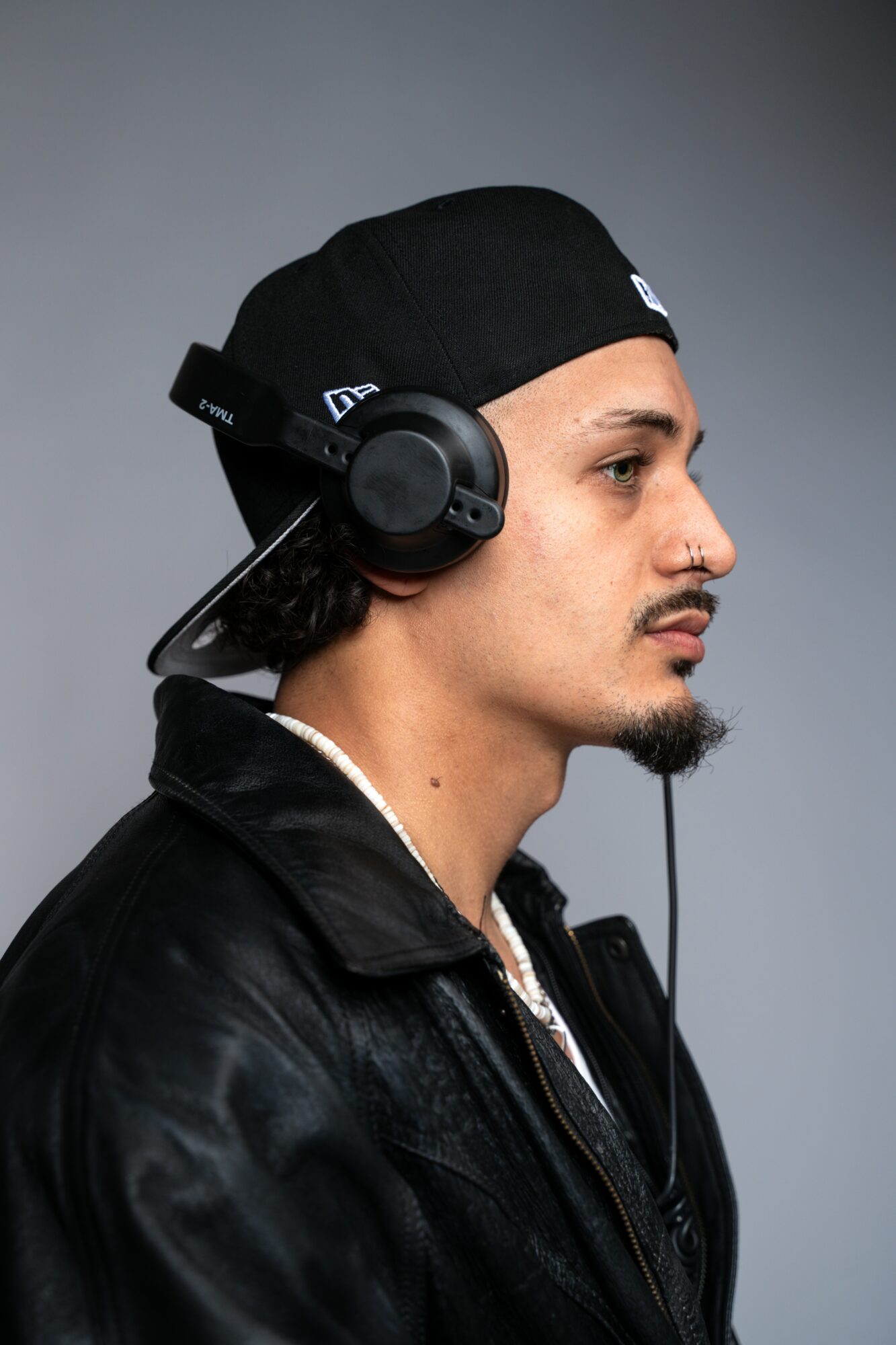 Side profile of a young man wearing a black cap, headphones, and a leather jacket against a gray background.