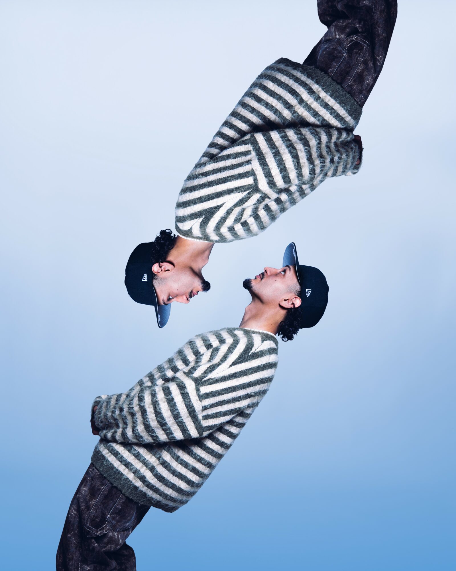 Two women face each other, touching foreheads, wearing striped sweaters and black hats, against a blue background.