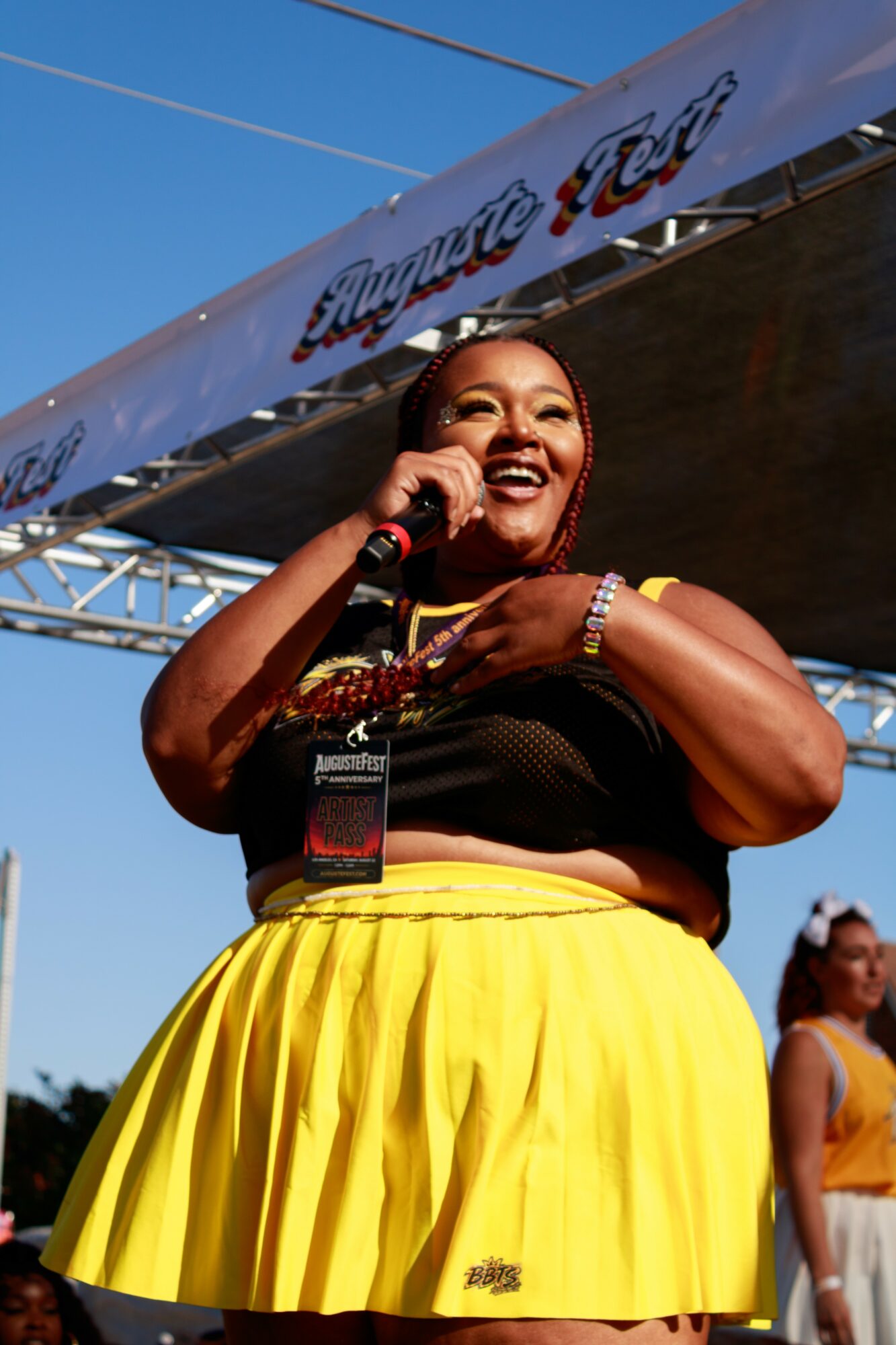 Smiling woman holding microphone on stage, wearing yellow skirt and black top, with a clear blue sky background.
