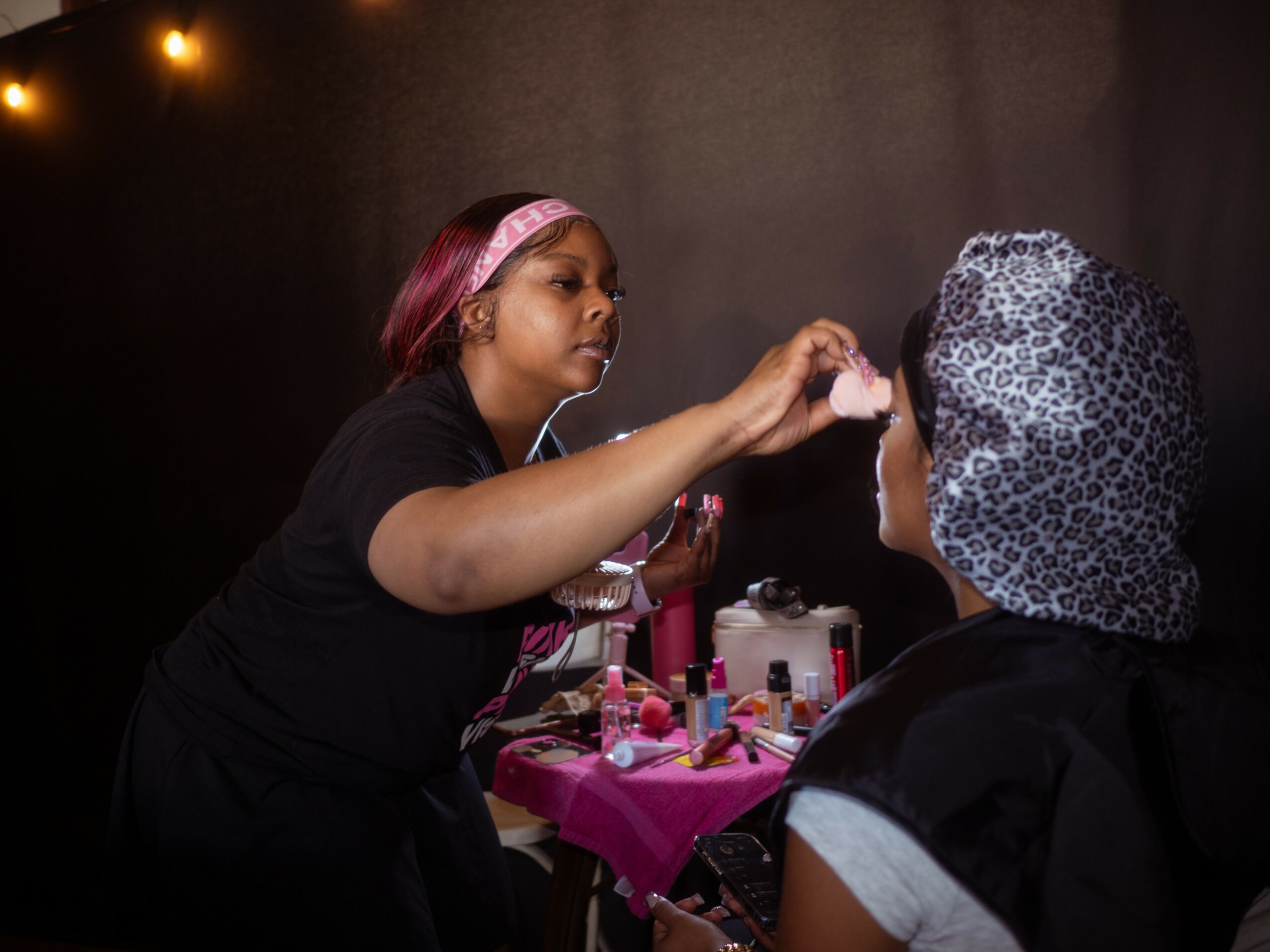 Makeup artist applying makeup to a person with a patterned headscarf, pink makeup tools on a table, dark background.