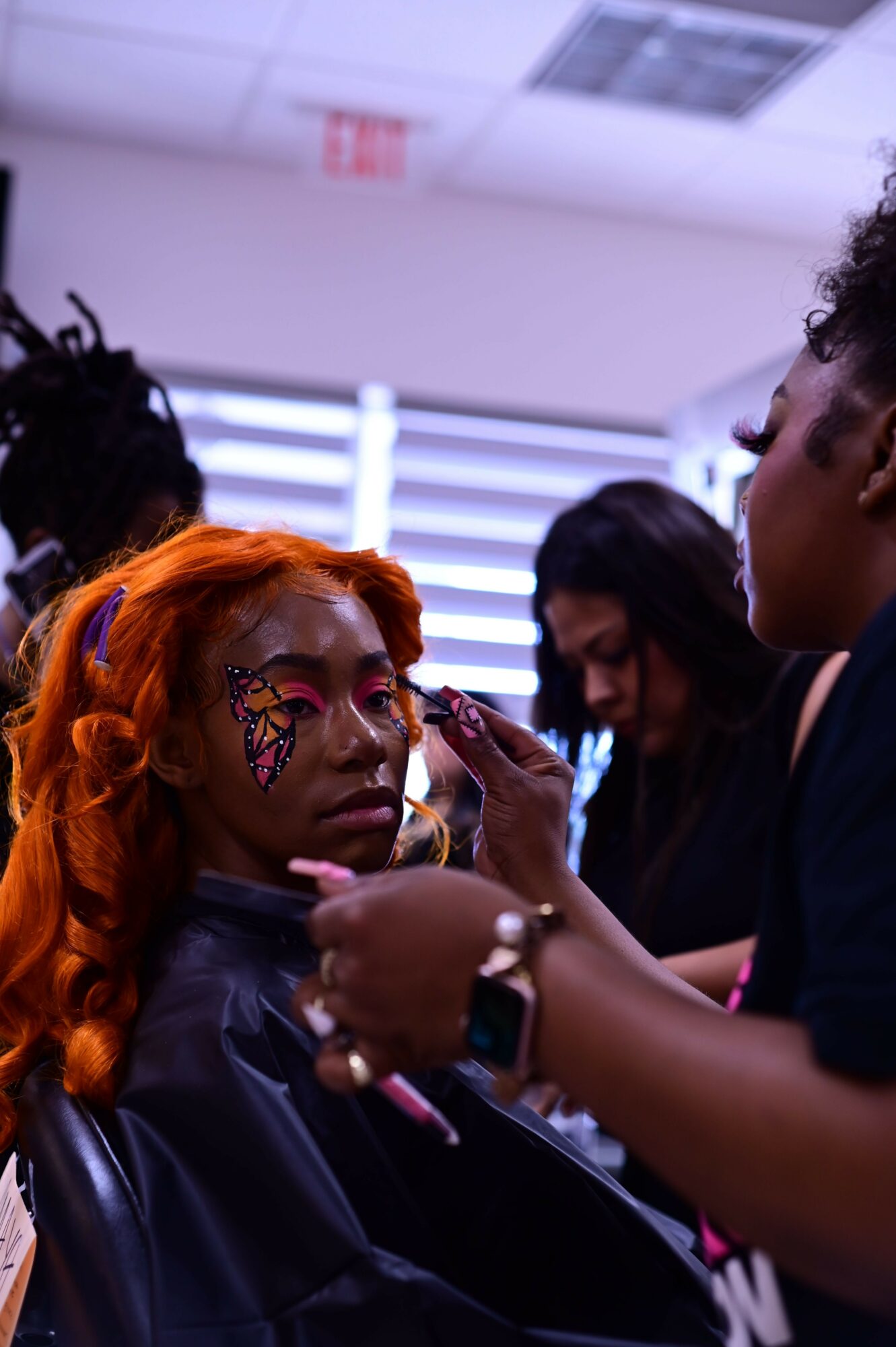 Person with orange hair and face paint receives makeup application from another person, indoors with vertical blinds in background.