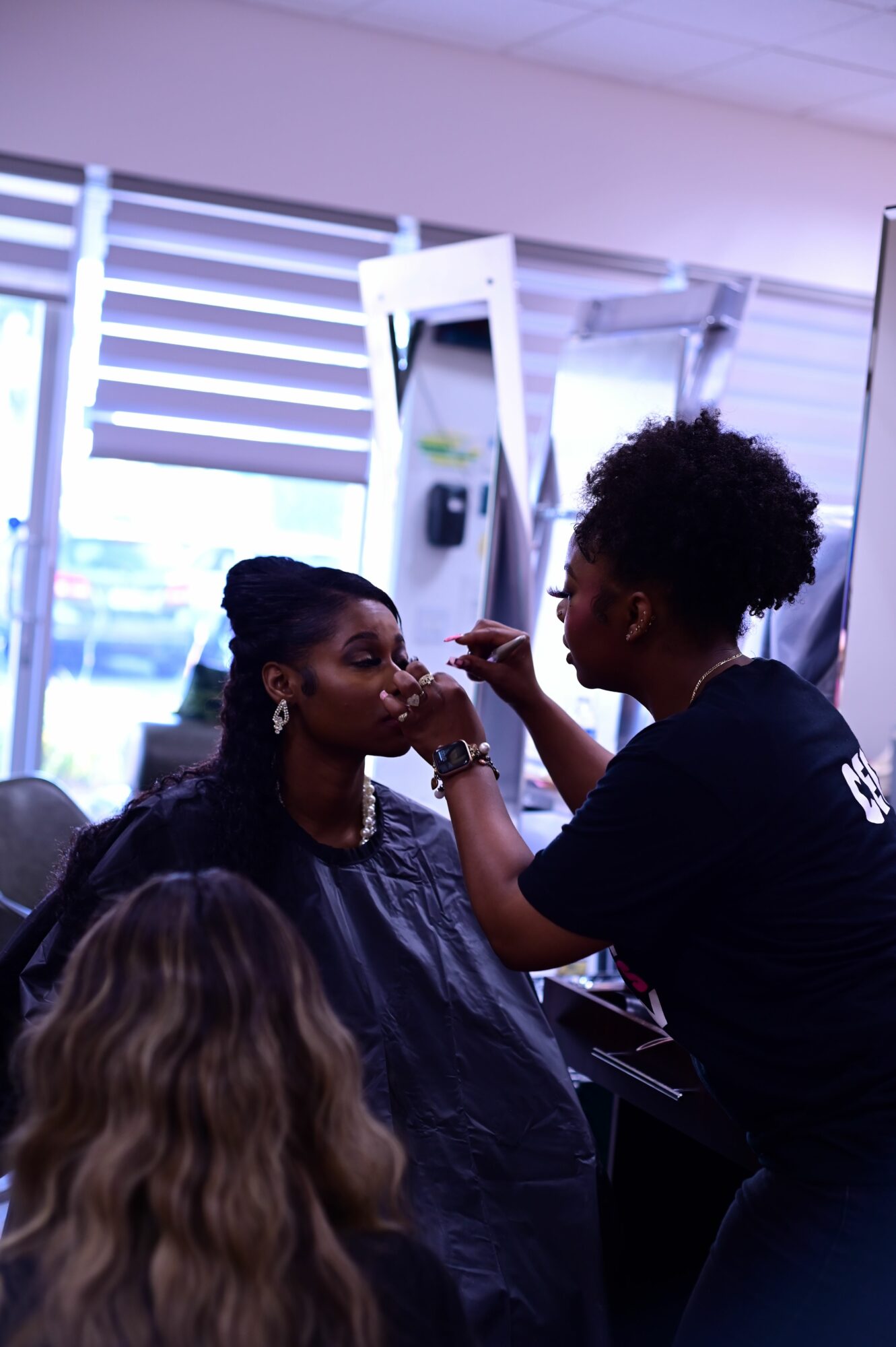 Two women in a salon, one styling the other's hair near a large window with blinds.