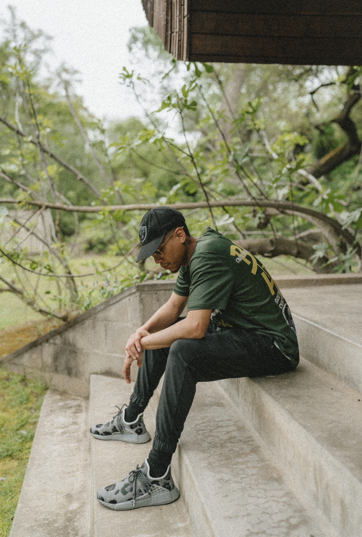 Person sitting on concrete steps outdoors, wearing a dark cap, green shirt, and gray sneakers, looking down.
