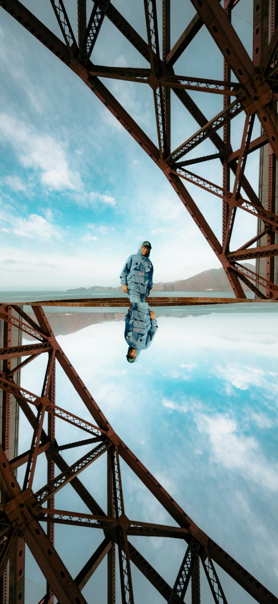 Person standing on a reflective surface under a large metal bridge structure with blue sky and clouds.