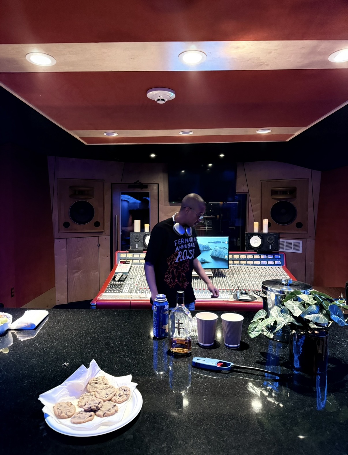 Person in a recording studio with audio equipment, mixing console, and snacks on a table, facing away from the camera.