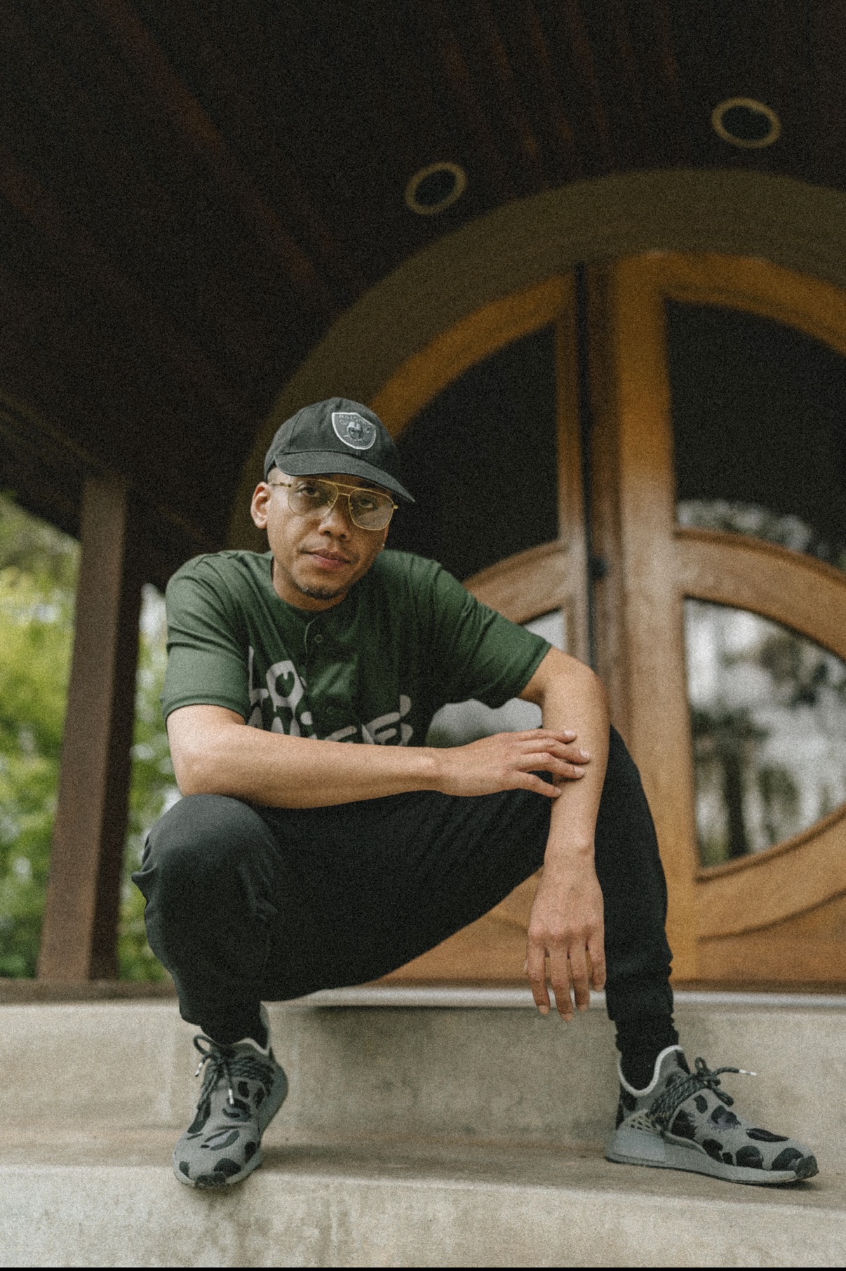 Person crouching on steps in front of a wooden door with circular window, wearing glasses, a cap, and athletic shoes.