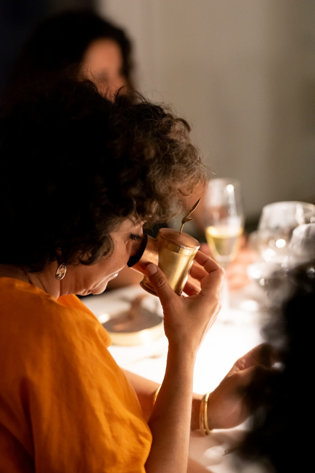 Person with curly hair holding a small cup, sitting at a table with drinks and tableware, in a social setting.