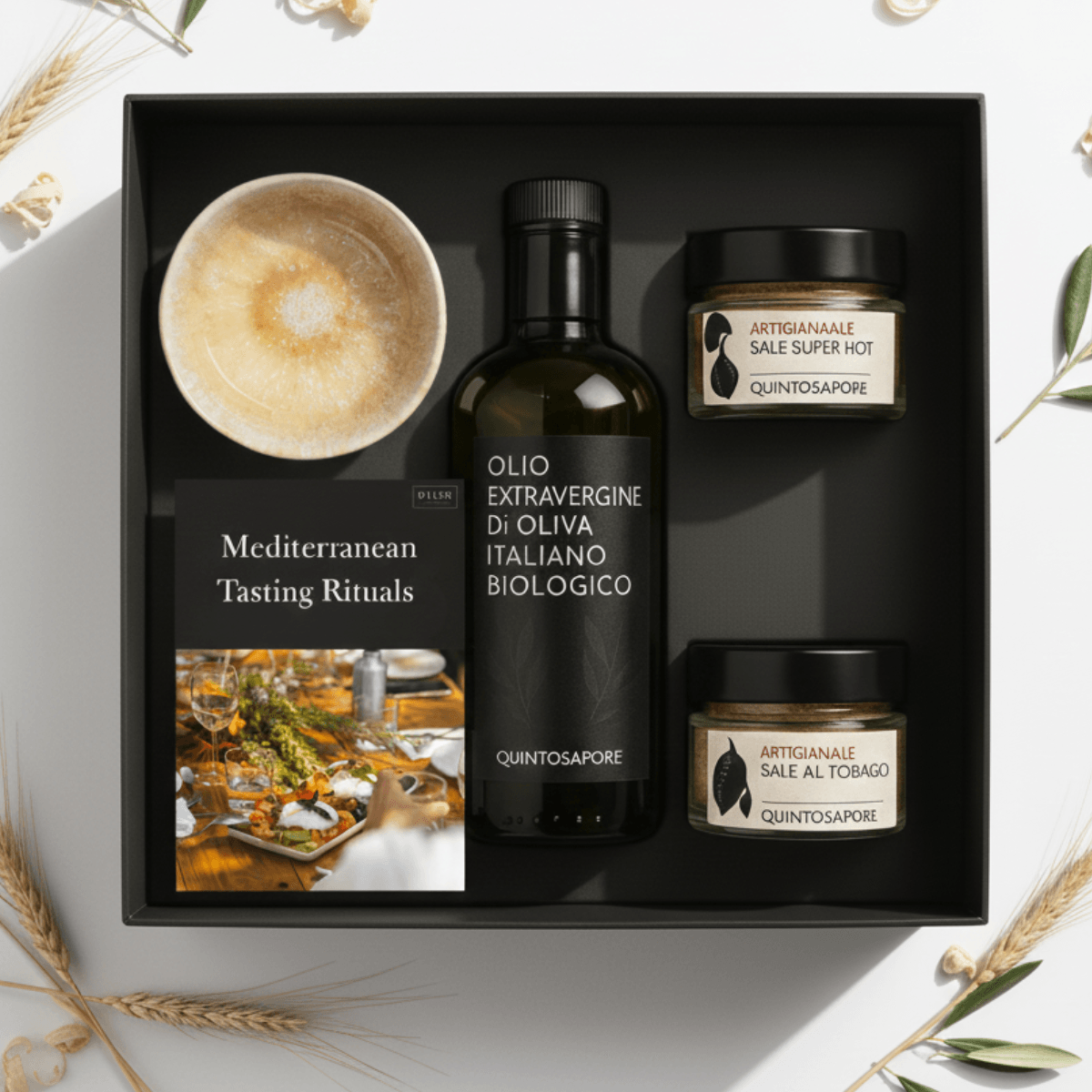 Black box containing olive oil and two jars, a cup, and a card with Mediterranean tasting rituals, on a white surface with plants.