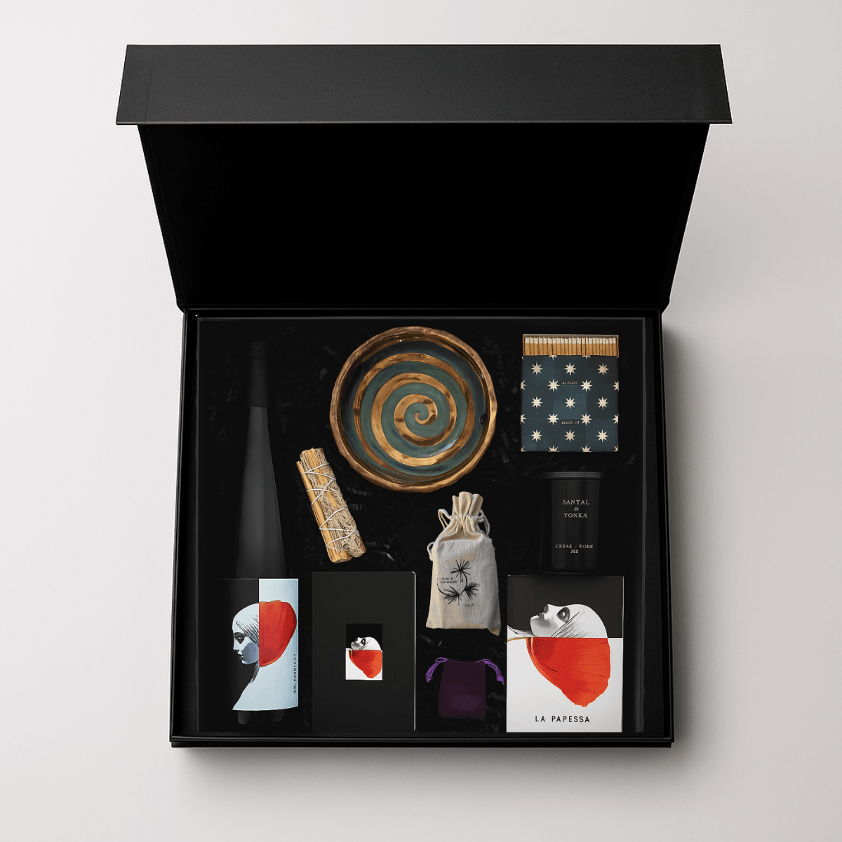 Open black box containing a spiral-patterned plate, a polka dot box, a small black box, a wrapped item, and various cards or tags.