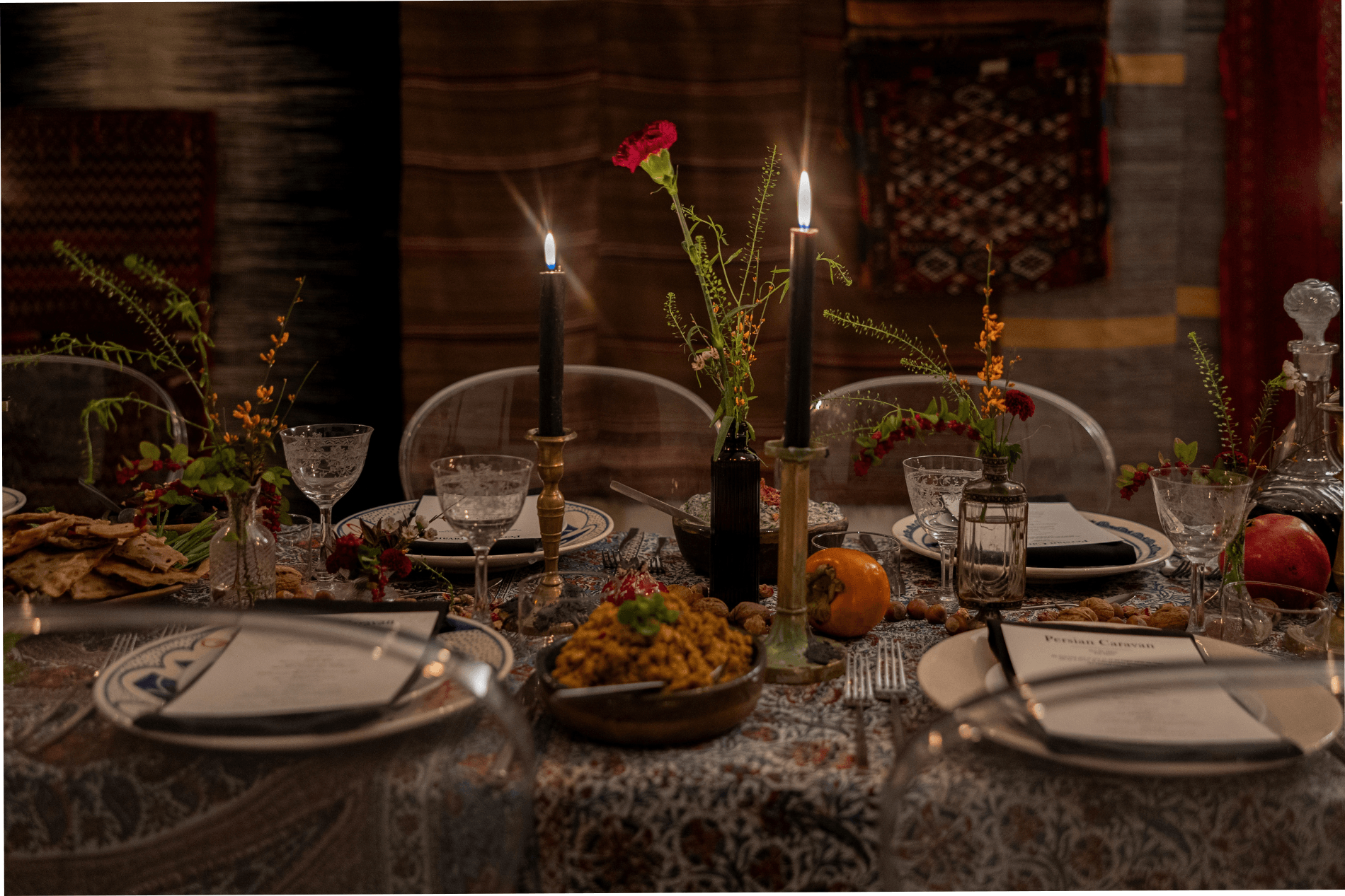 Table set with candles, flowers, plates, glasses, and food, in a dimly lit room with patterned decor.