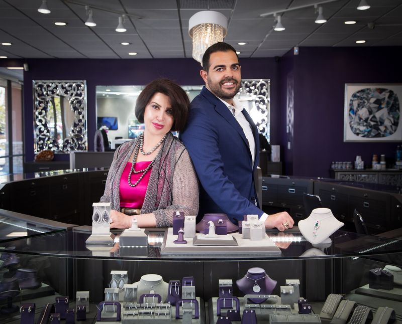 Two people, a woman and a man, stand behind a jewelry display counter in a store, smiling. The store has mirrors and artwork on the walls.
