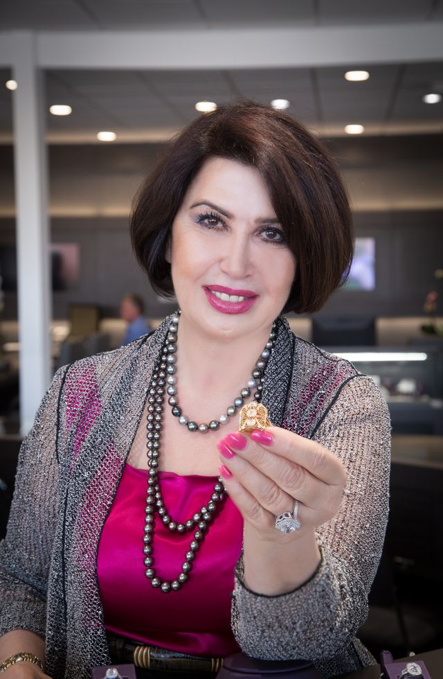 Woman with short dark hair smiling, holding a ring, wearing a pink top and layered necklaces, in an indoor setting.