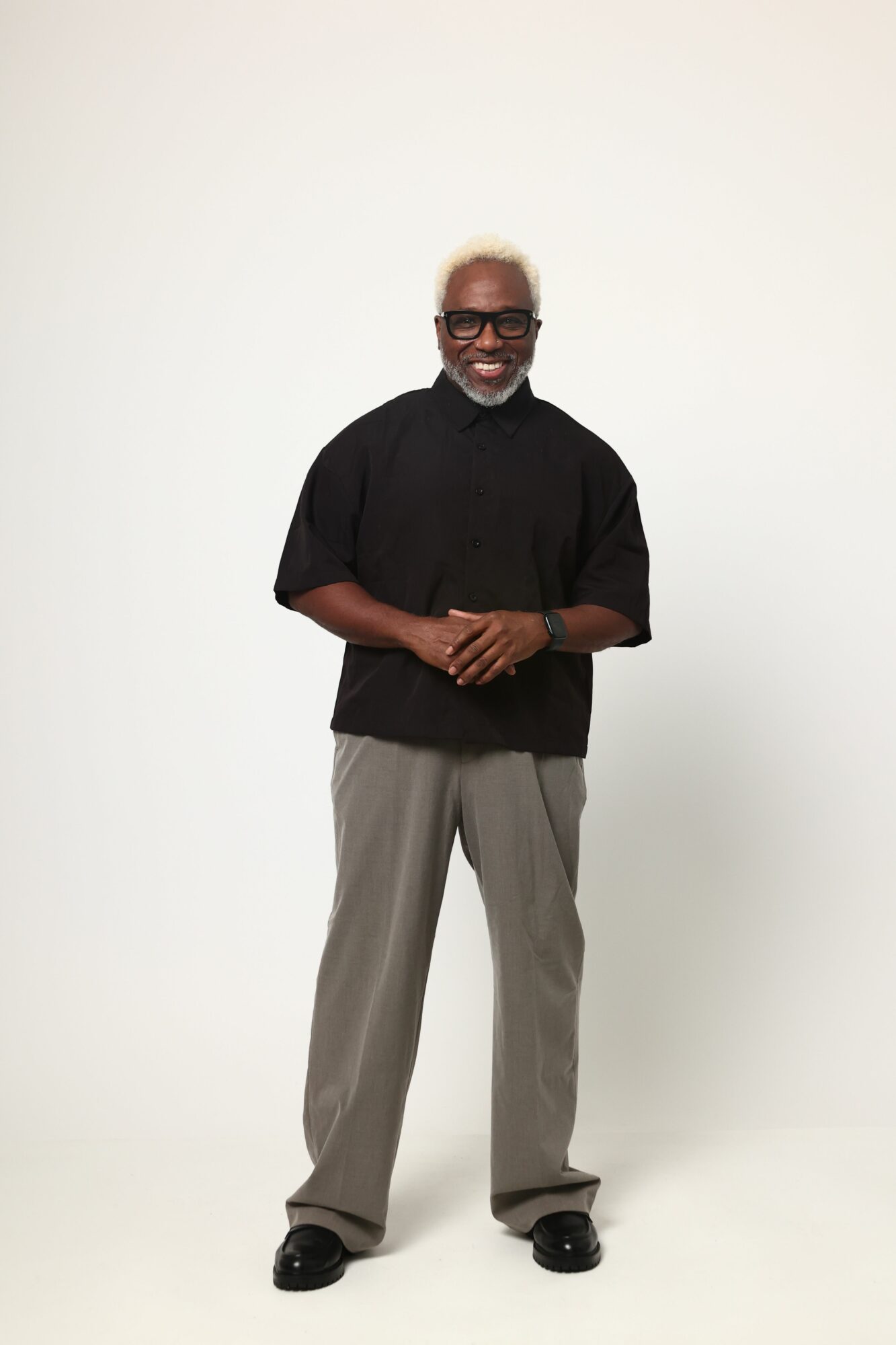 Man standing against a white wall, smiling, wearing glasses, a black shirt, gray pants, and black shoes.