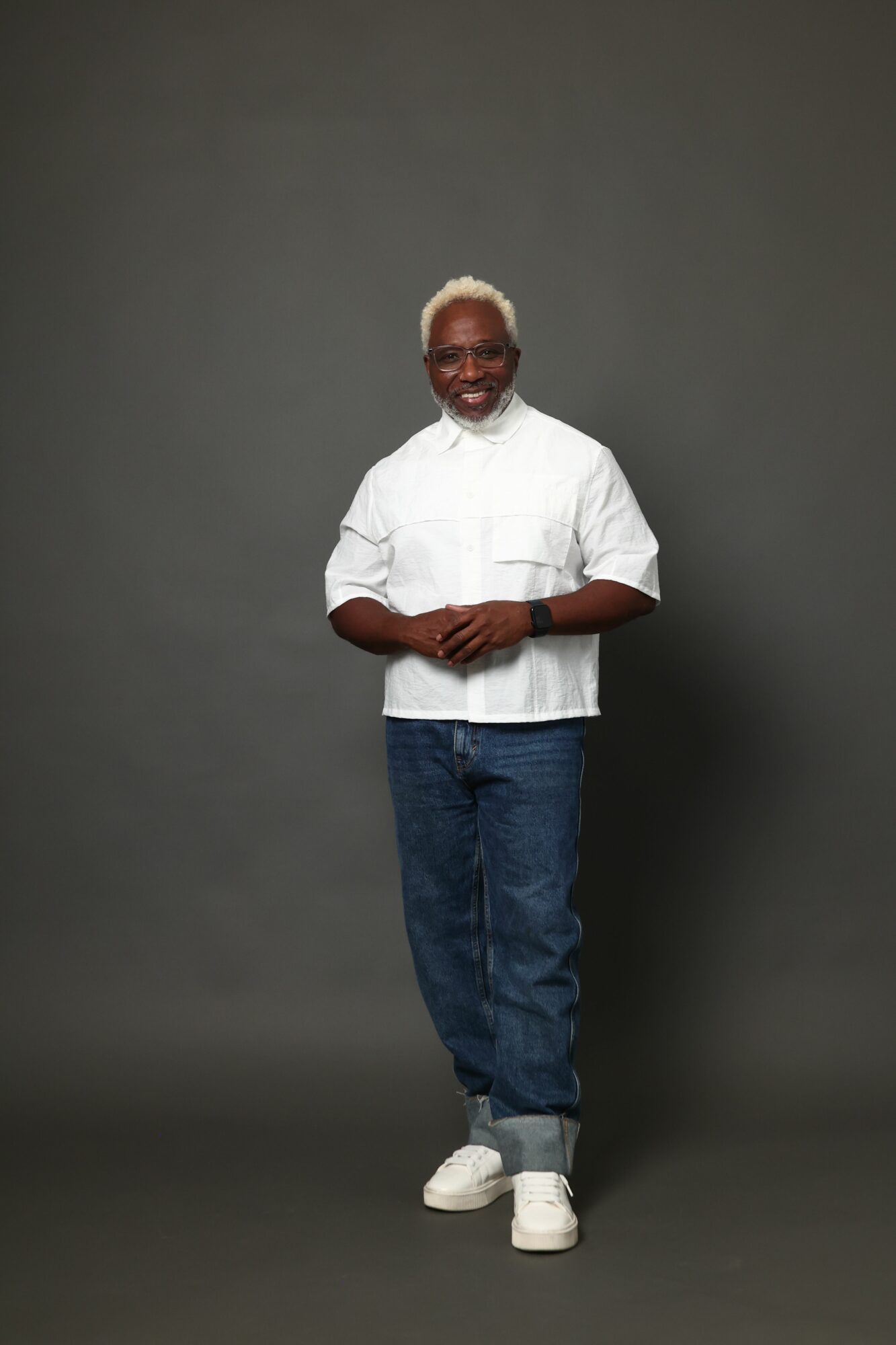 Older man with white hair wearing a white shirt, jeans, and white shoes, standing against a dark background.