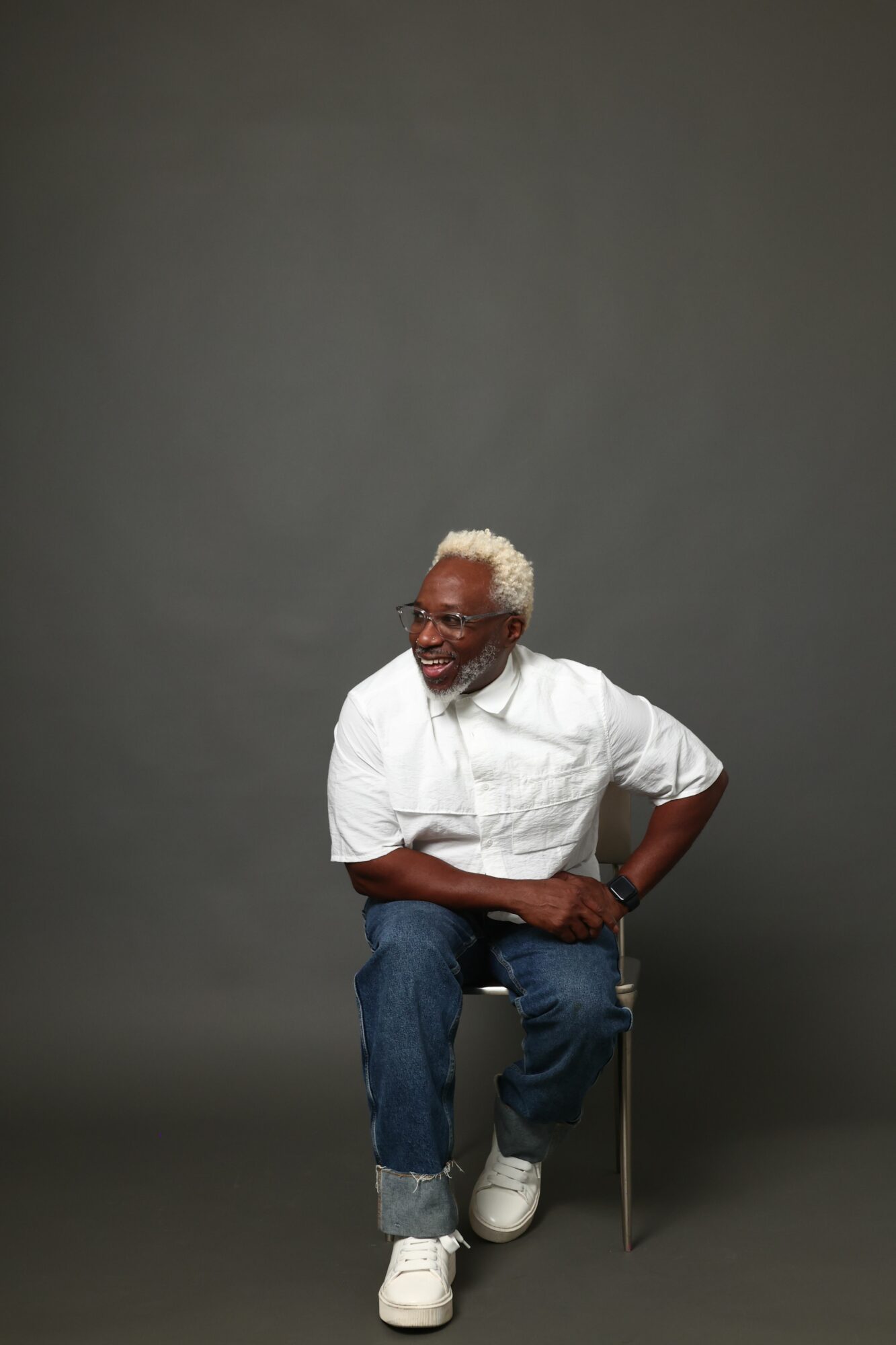 Elderly person with white hair and glasses, wearing a white shirt and jeans, holding a cane, standing against a gray background.