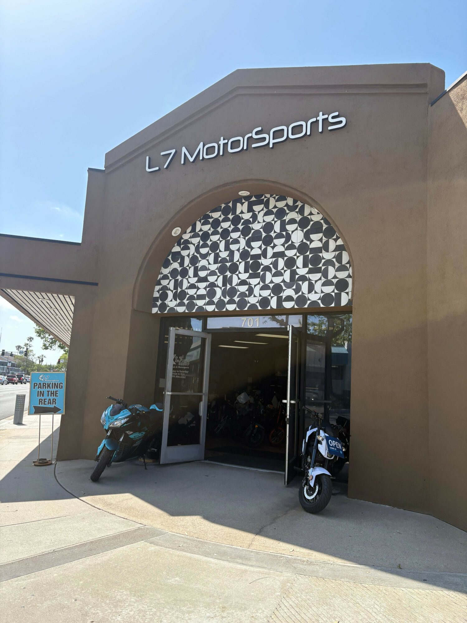 Sports store entrance with motorcycles inside, sign reads 'Motorsports', archway with patterned design, glass door, concrete sidewalk.