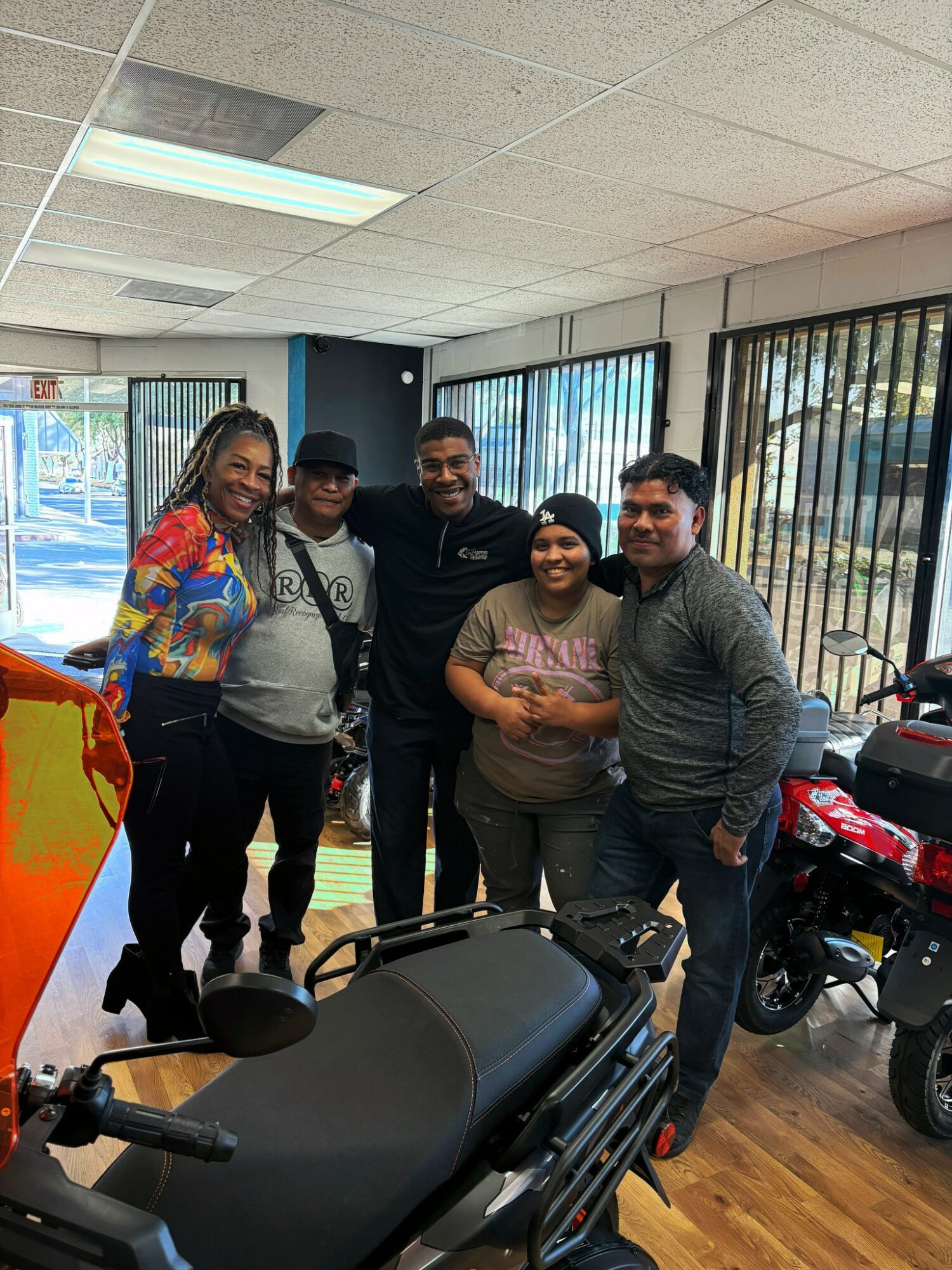 Group of five people standing indoors, smiling, with motorcycles and large windows behind them.